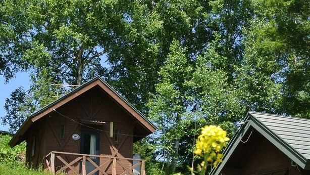 A rustic wooden cabin with a porch, surrounded by lush greenery and yellow flowers. - Pension Megumiyuki (Biei)