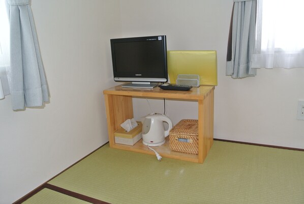 A small room with a wooden desk, a black monitor, a yellow lamp, a white kettle, and a woven basket. - Pension Megumiyuki (Biei)