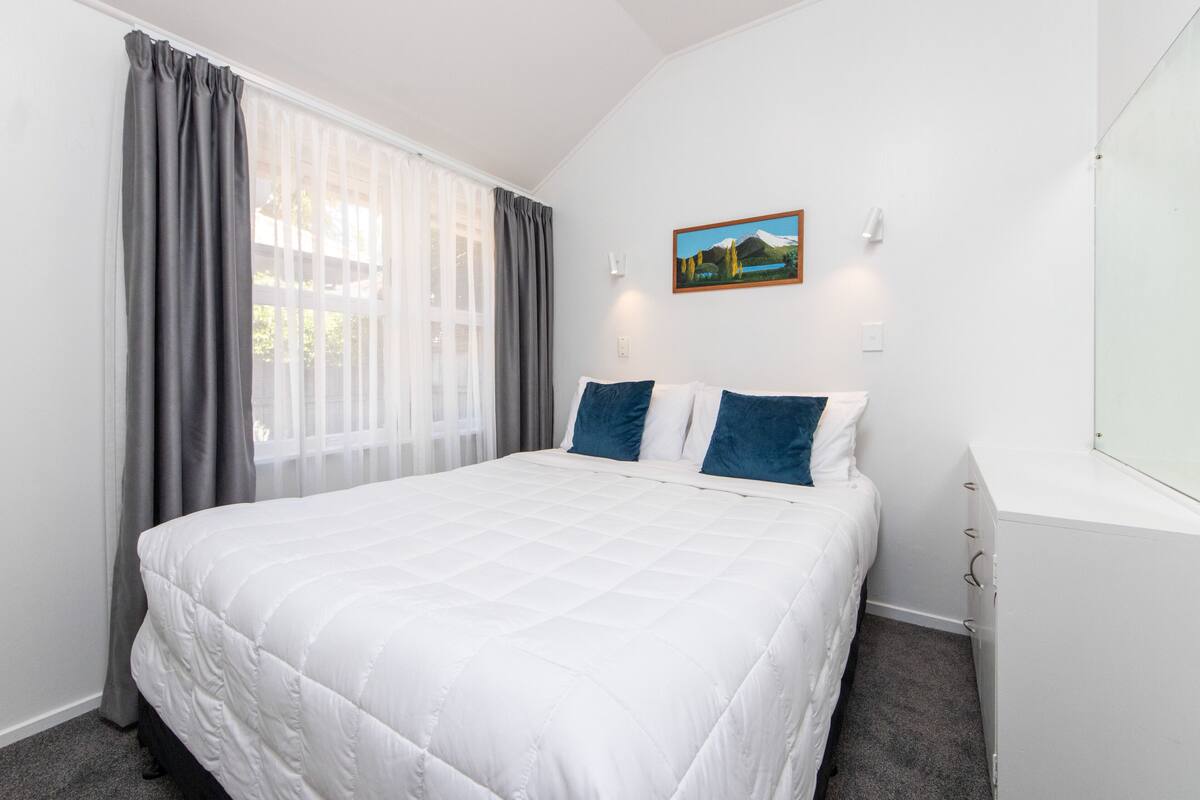 A neatly made bed with a white duvet, grey curtains, and a painting on the wall.