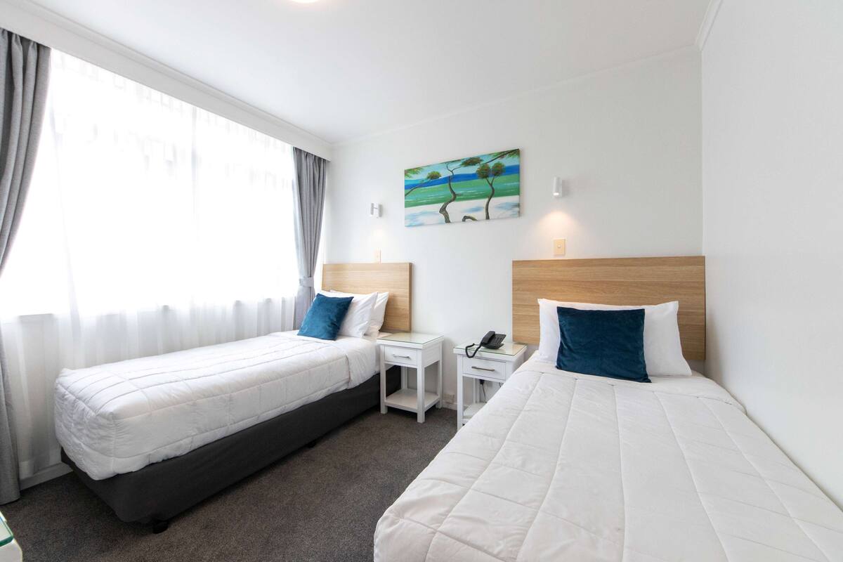 A hotel room with two single beds, a small bedside table with a phone, a painting on the wall, and a window with curtains.