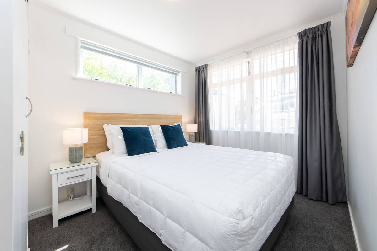 A hotel room with a large bed, a bedside table with a lamp, a window with curtains, and a view of the outdoors.