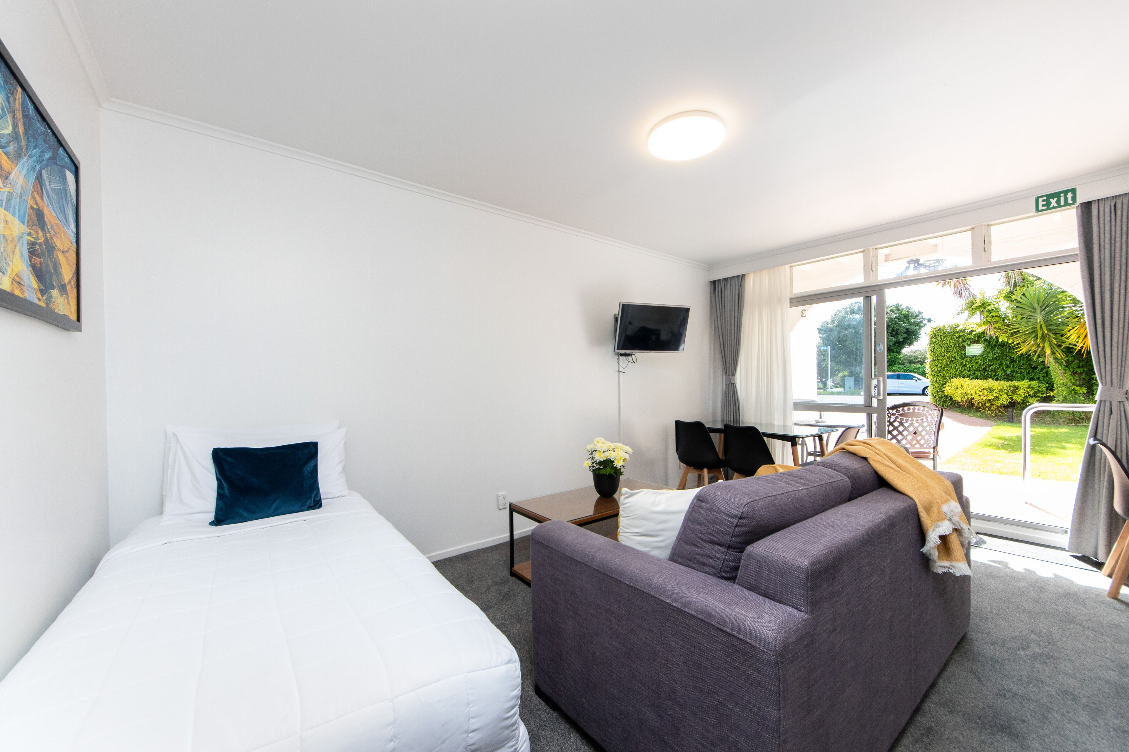 Et hotelværelse med seng, sofa, et lille bord med blomster i en vase, et fjernsyn ophængt på væggen og udsigt til det fri gennem et stort vindue.