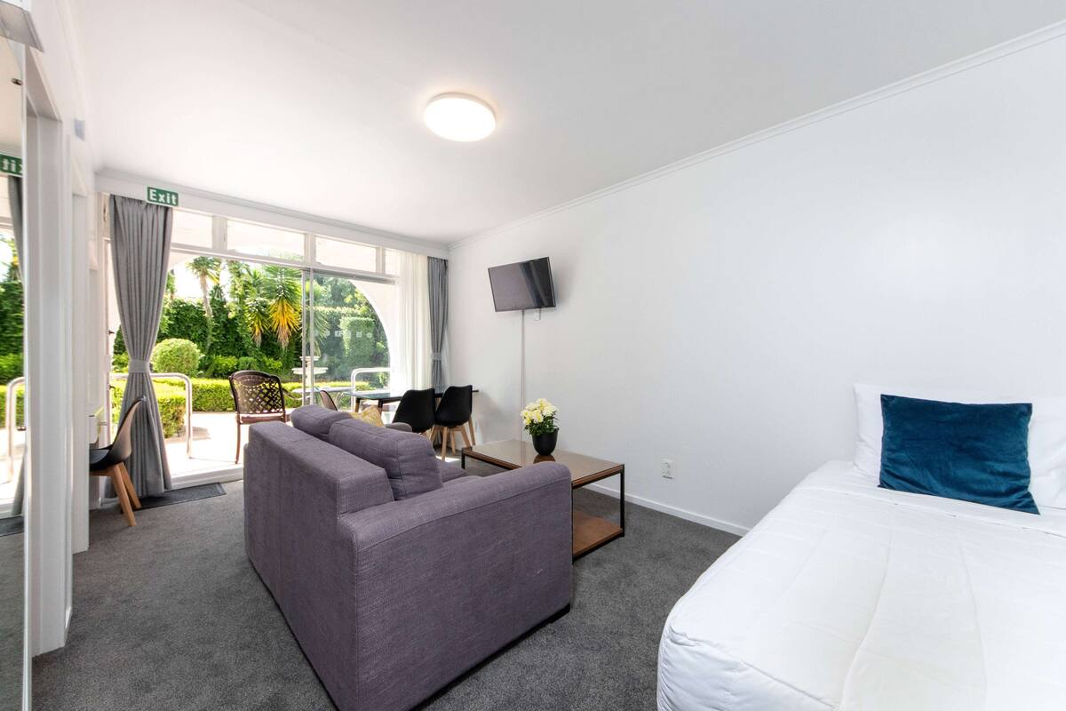 A hotel room with a sofa, a bed, a small table with a flower, and a TV on the wall.