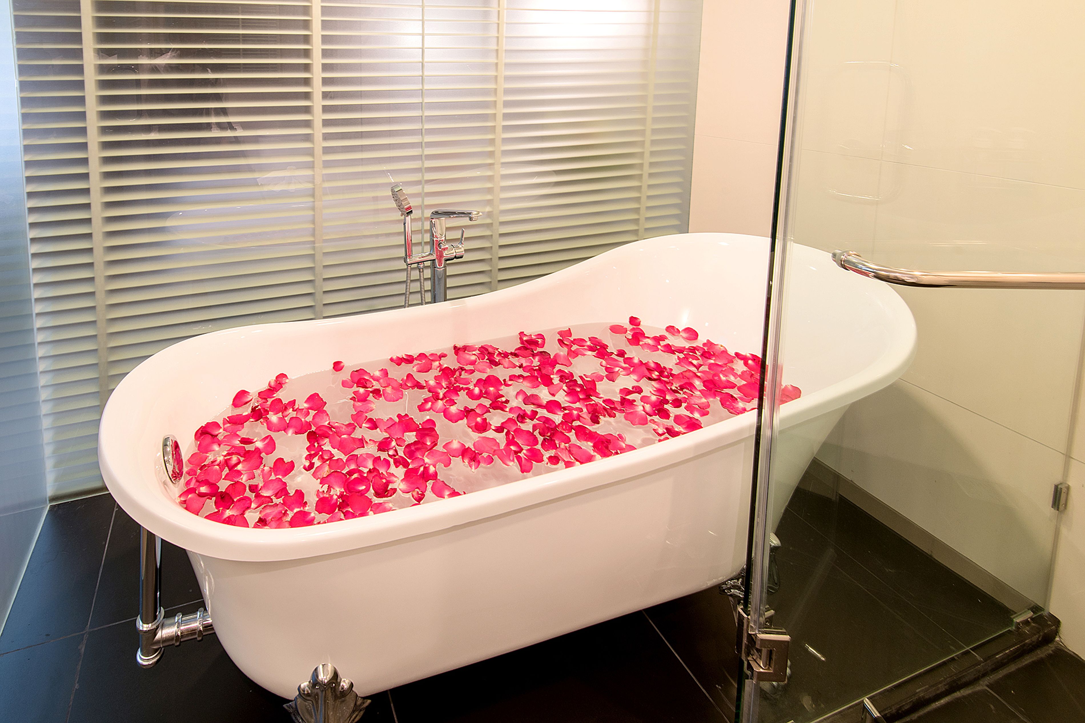 Una bañera blanca independiente llena de pétalos de rosa, cabina de ducha de vidrio y una ventana con persianas.