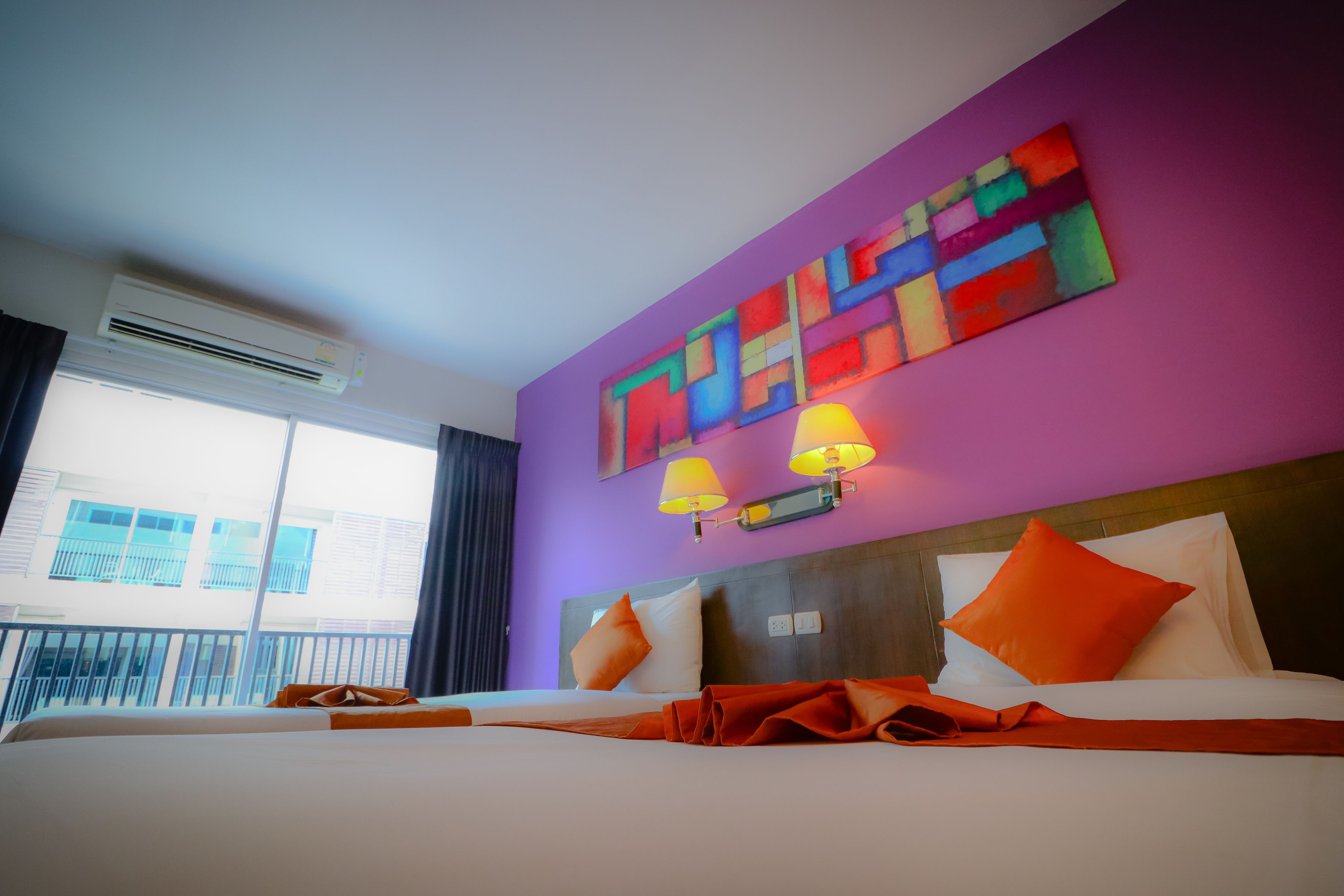 A hotel room with a large bed, a colorful abstract painting on the wall, and a balcony with a view of buildings.