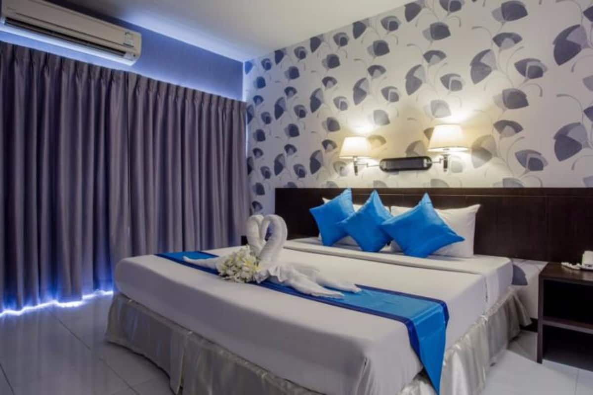 Una habitación de hotel con cama, mesita de noche, equipo de aire acondicionado, pared con diseño de hojas y ventana con cortinas.