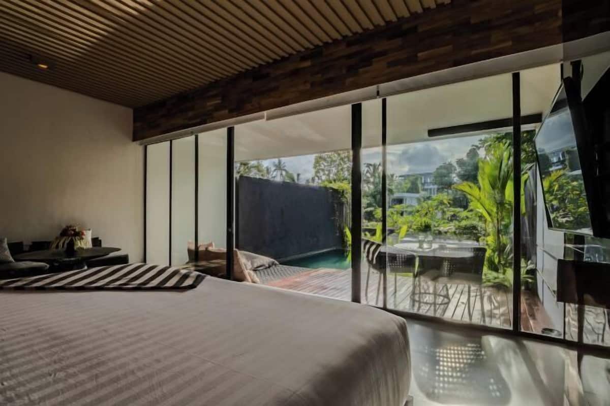 A bedroom with a large bed, a seating area with a sofa and chairs, and a view of an outdoor deck with a table and chairs.