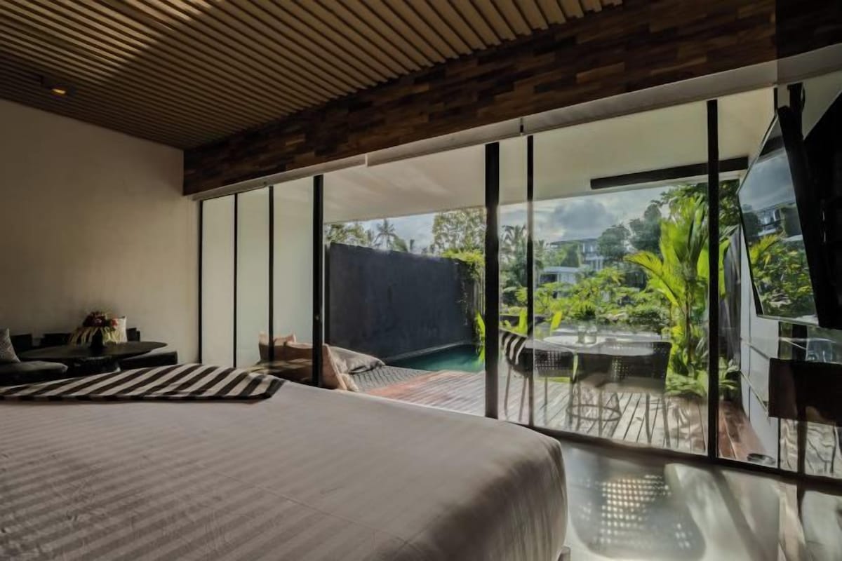 A bedroom with a large bed, a seating area with a sofa and chairs, and a view of an outdoor deck with a table and chairs.
