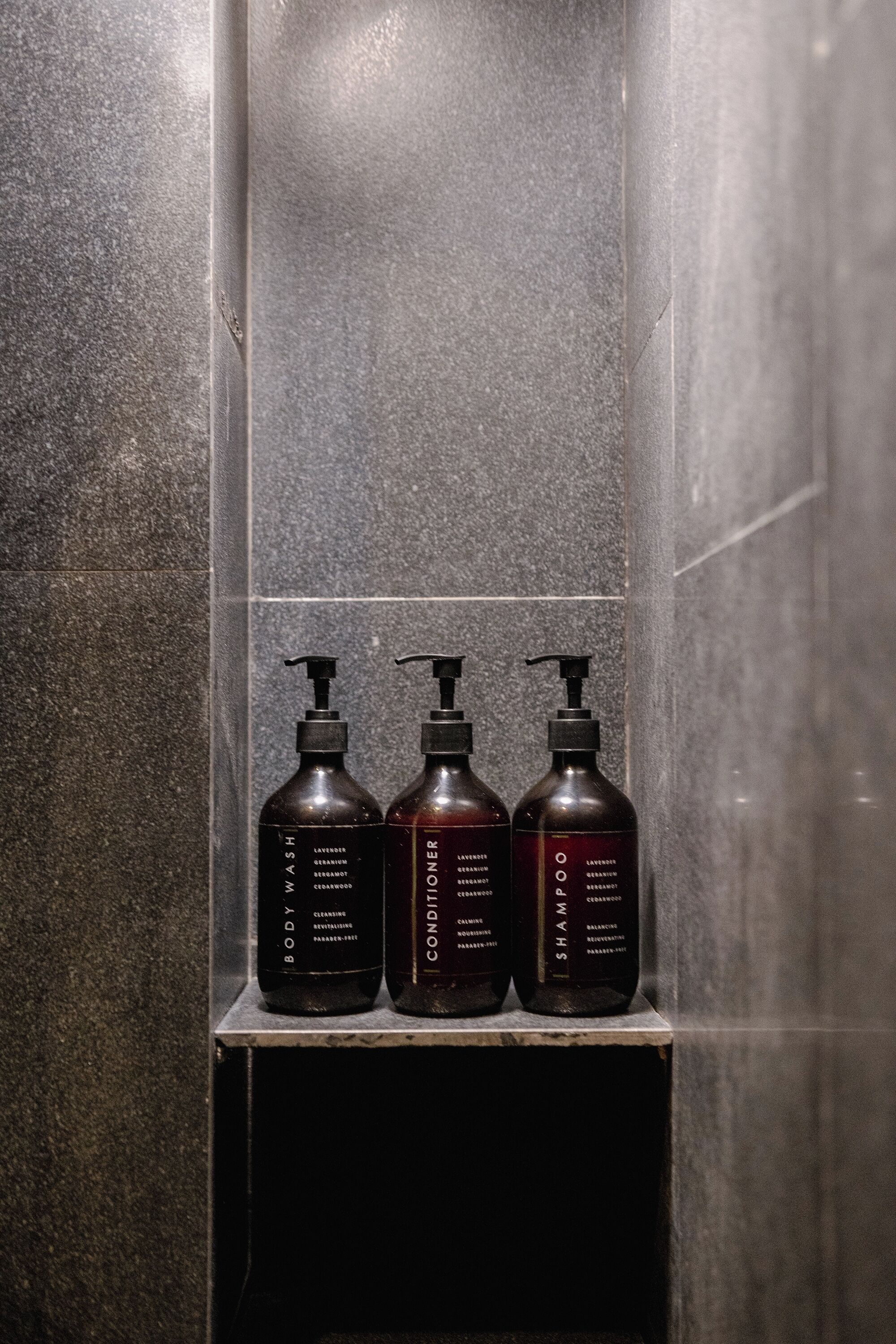 Three bottles of body wash and conditioner on a shelf in a bathroom with a tiled wall.