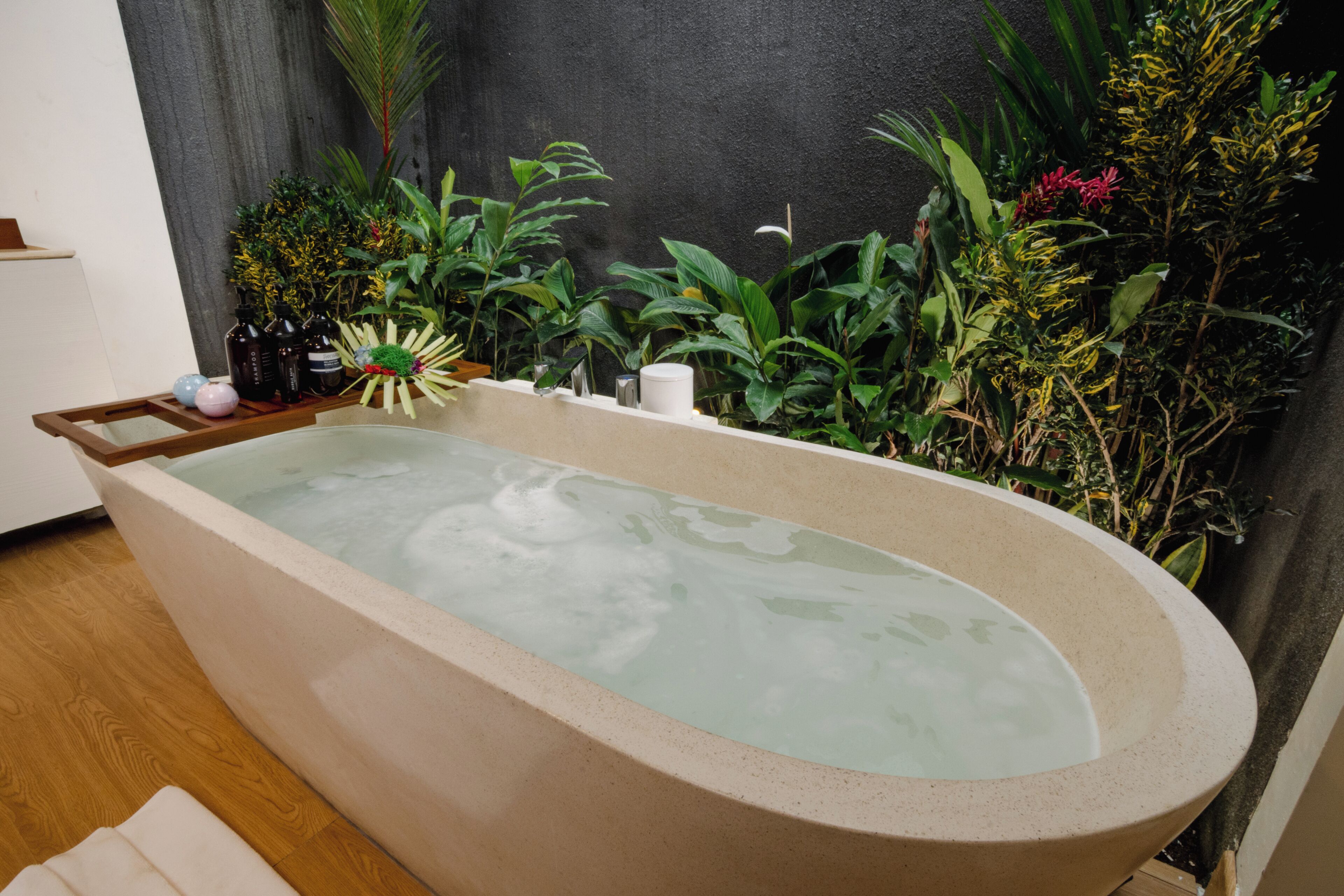 A bathtub with a wooden tray, surrounded by various plants and a dark wall.