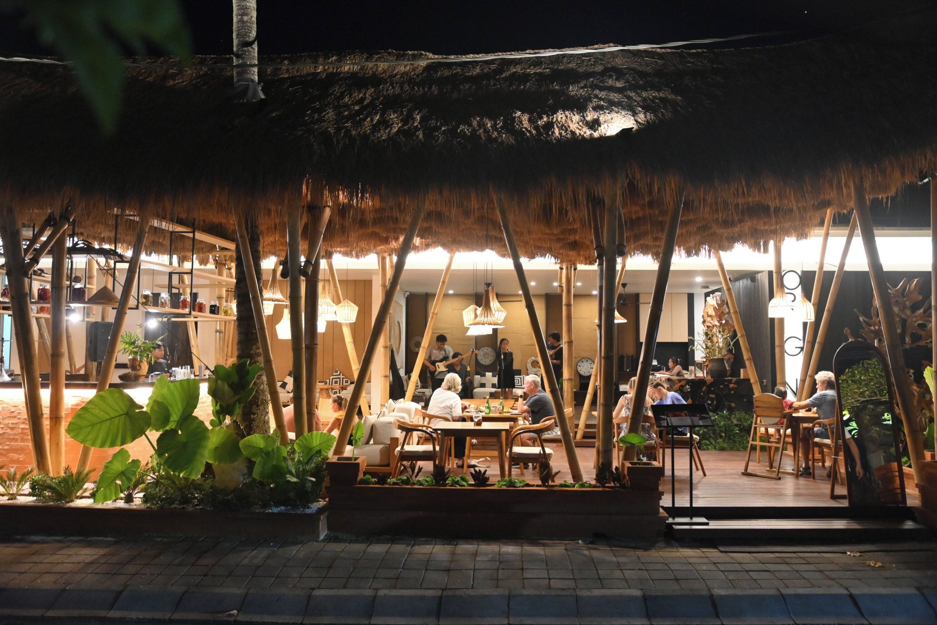 Un área de comedor al aire libre con techos de paja, mesas y sillas de madera, rodeada de plantas en macetas.