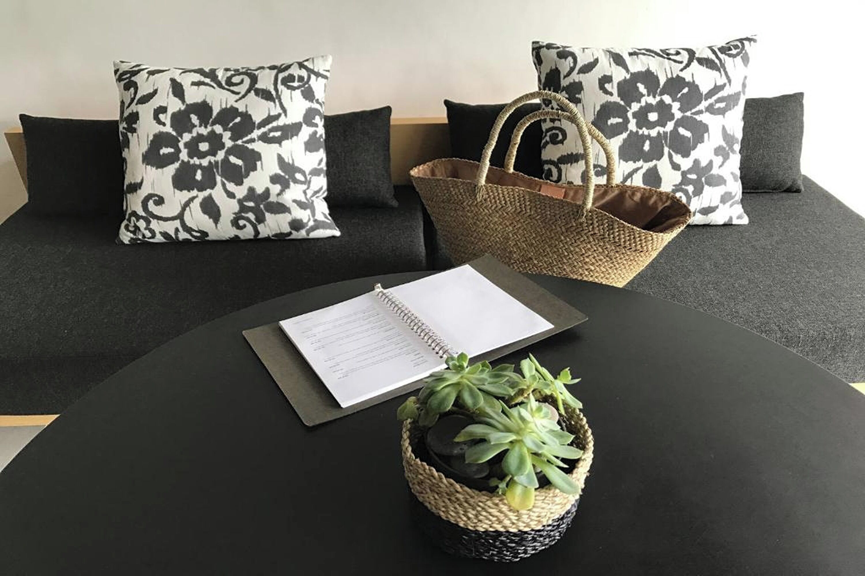 A black sofa with patterned pillows, a woven basket, a notebook, and a small potted plant on a black table.