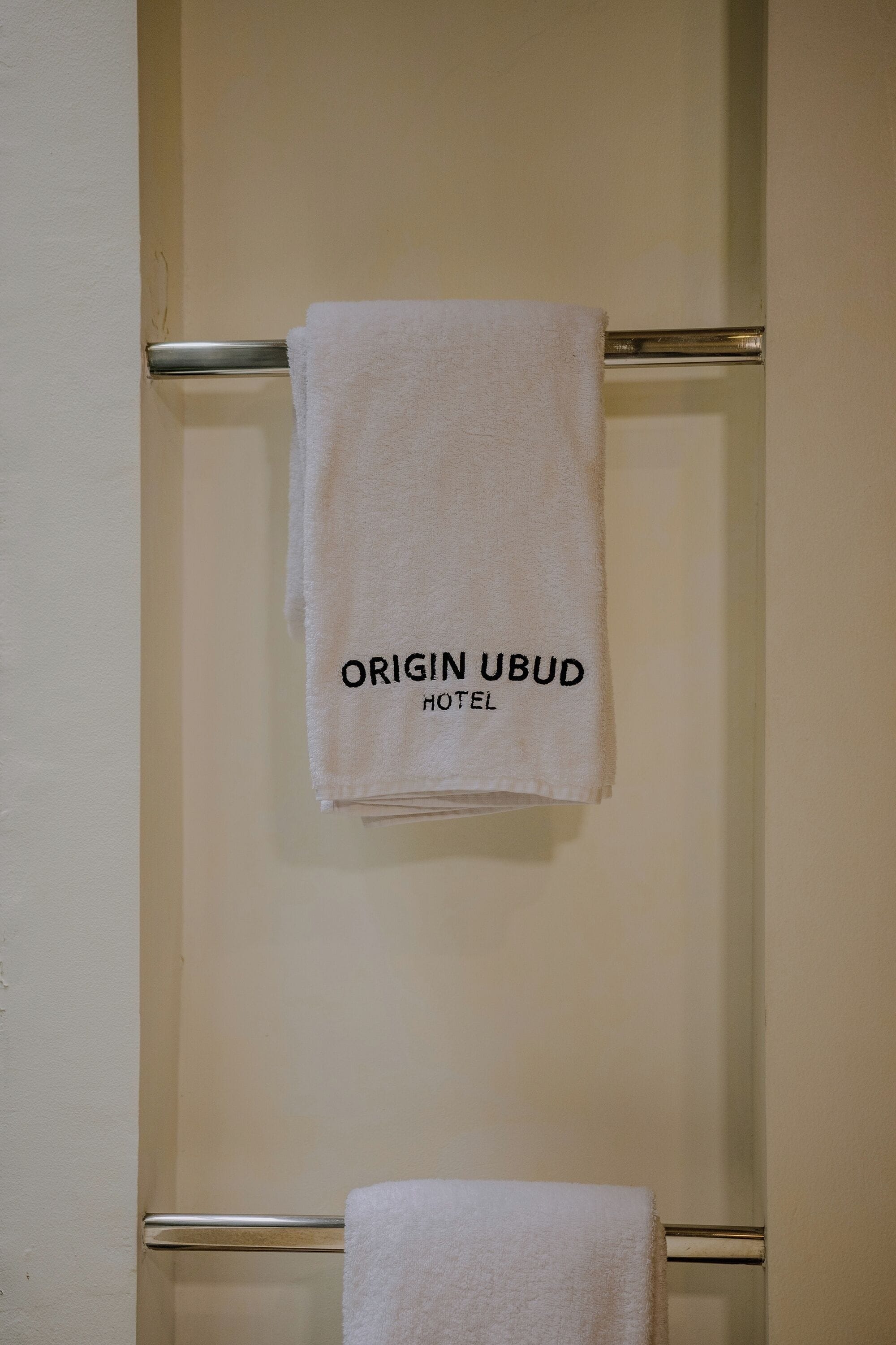 A white towel hanging on a metal towel rack with the text 'ORIGIN UBD HOTEL' on it.