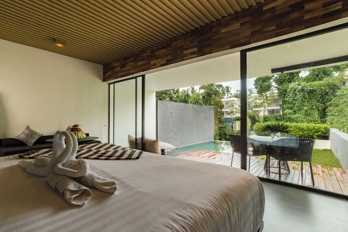 A modern hotel room with a large bed, a view of the outdoors, and a small pool area.