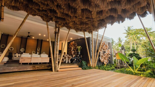 Un interior moderno con terraza de madera, techo de paja y zona de estar con sofás y mesa de comedor.