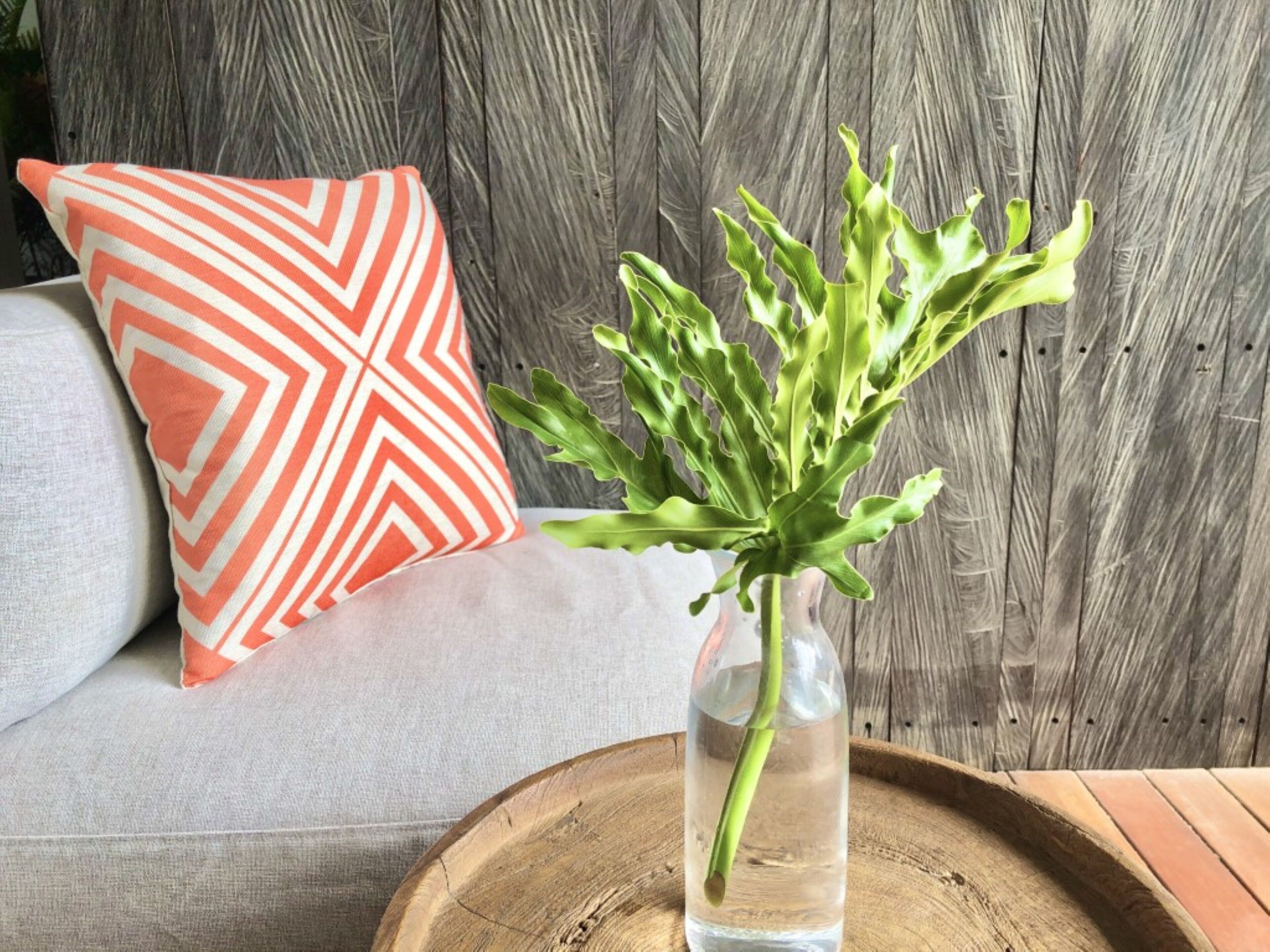 A vase with green leaves on a wooden tray, a gray sofa, and an orange and white pillow.