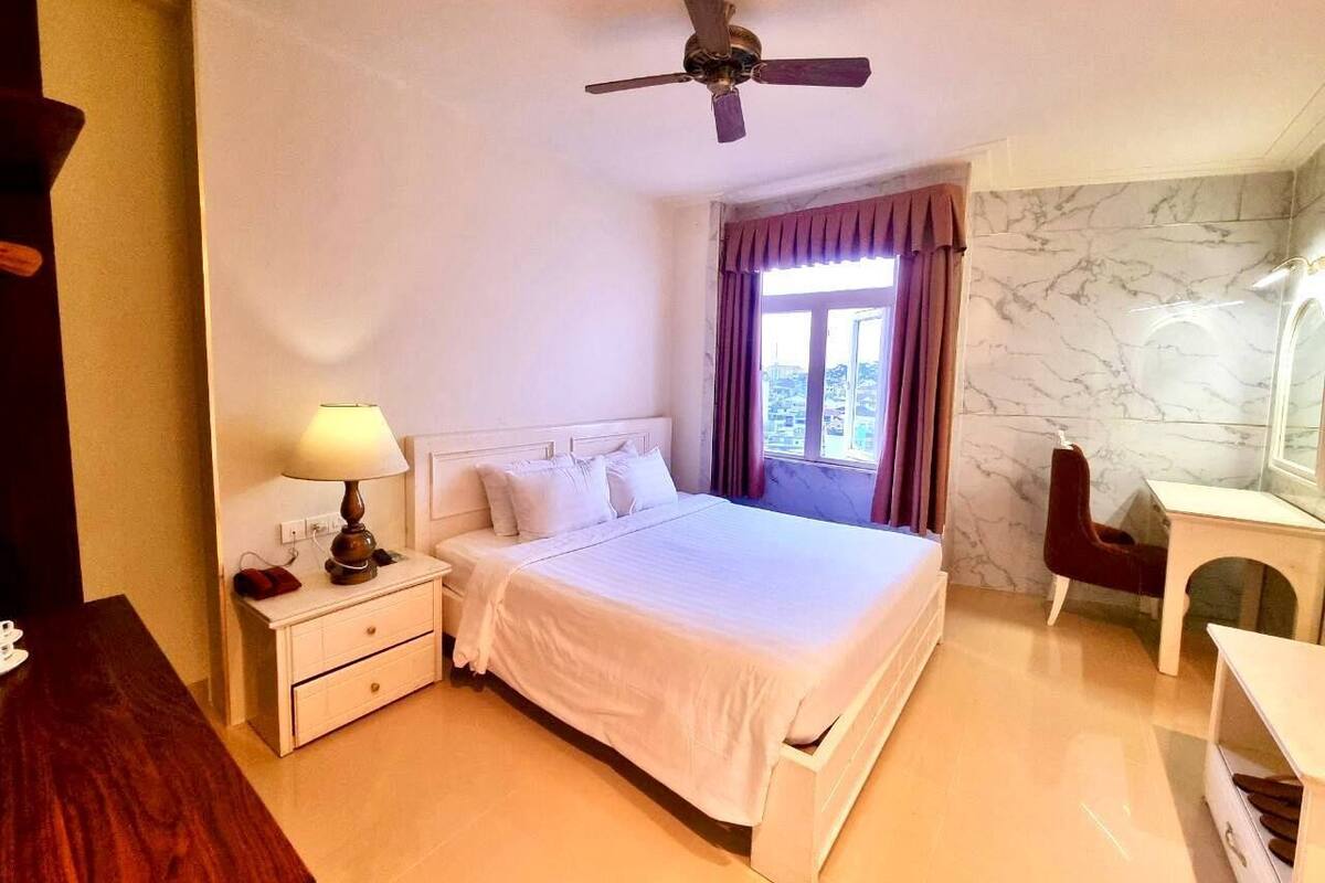 A hotel room with a bed, bedside table, lamp, desk, chair, and a ceiling fan.