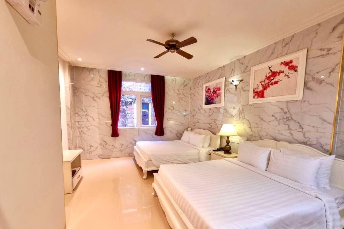 A hotel room with two beds, marble walls, a ceiling fan, a window with red curtains, and artwork on the wall.