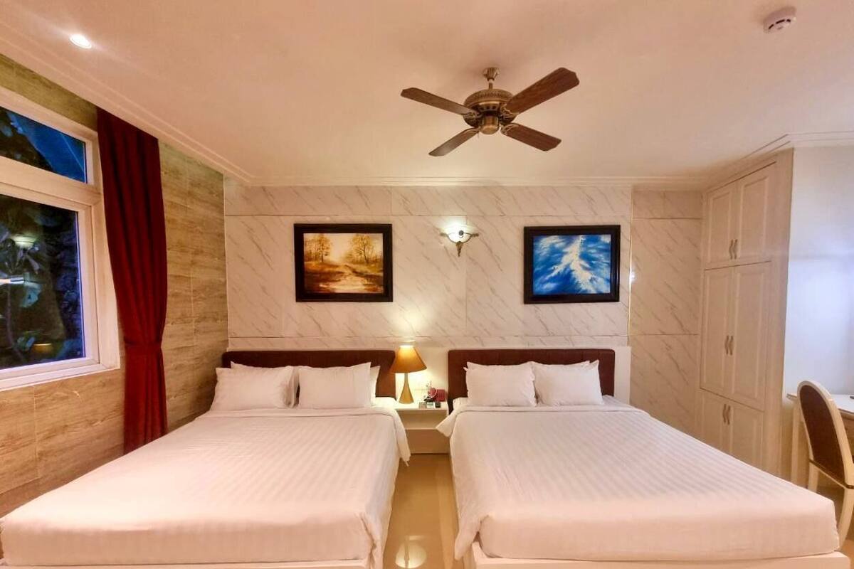 A hotel room with two beds, a ceiling fan, a nightstand with a lamp, and a window with red curtains.