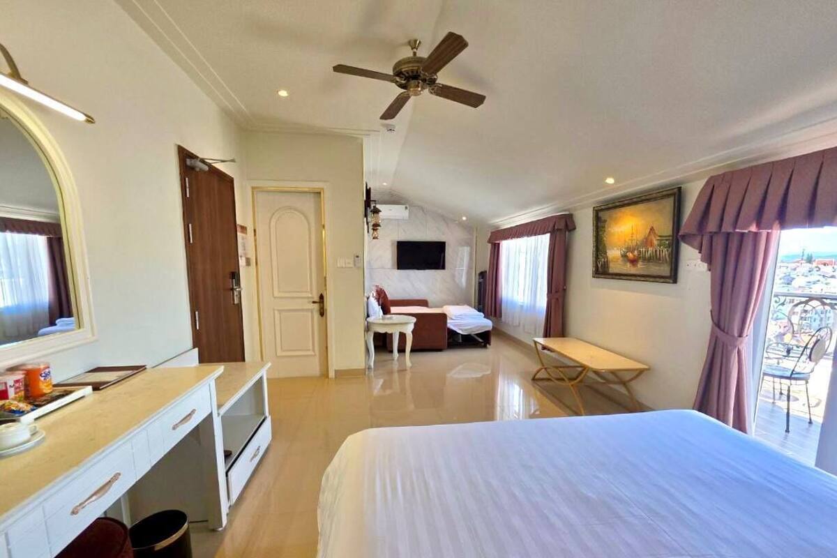 A hotel room with a bed, a desk, a chair, a small table, a ceiling fan, a TV, and a balcony with a view.