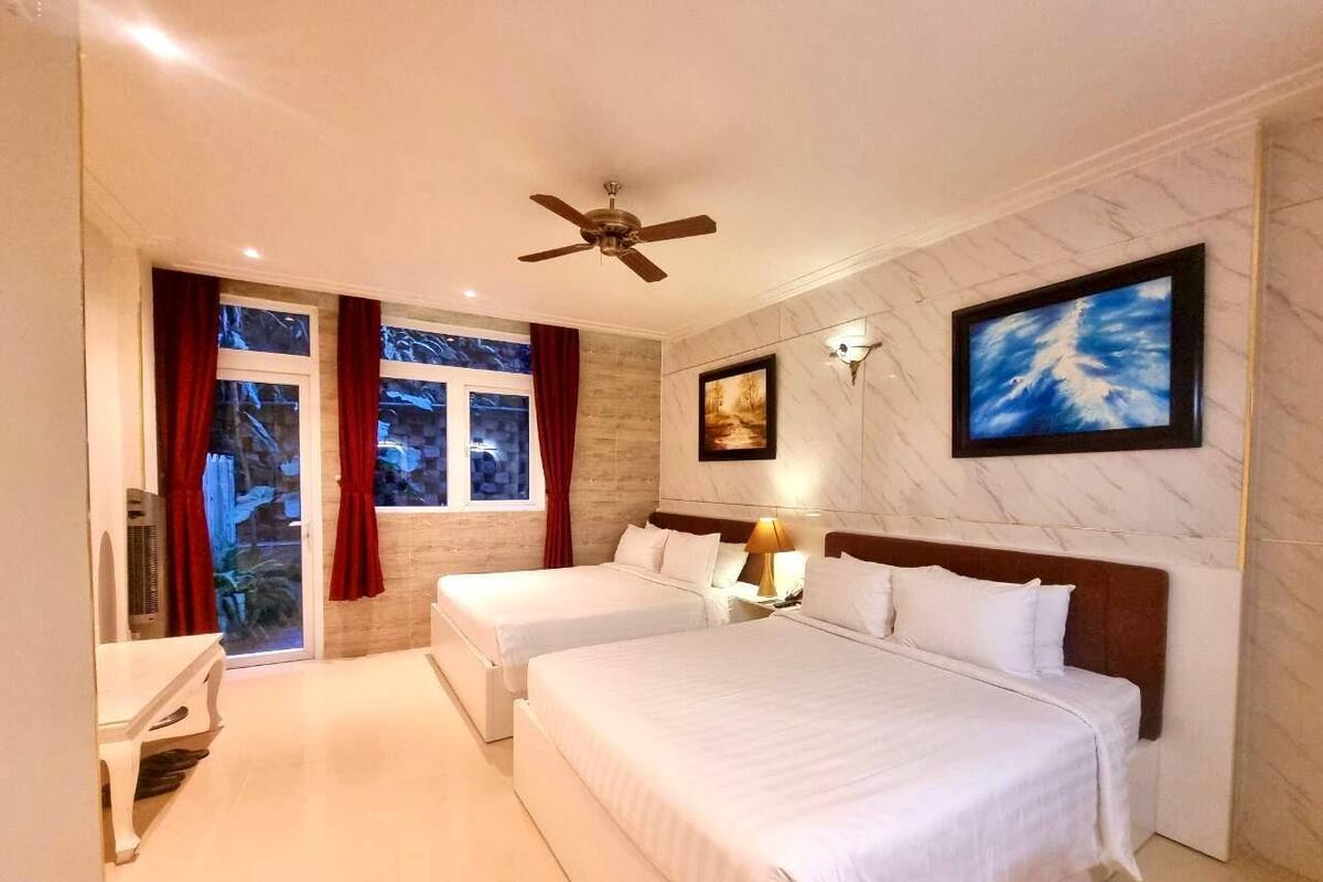 A hotel room with two beds, a ceiling fan, a large window with red curtains, and a marble wall.