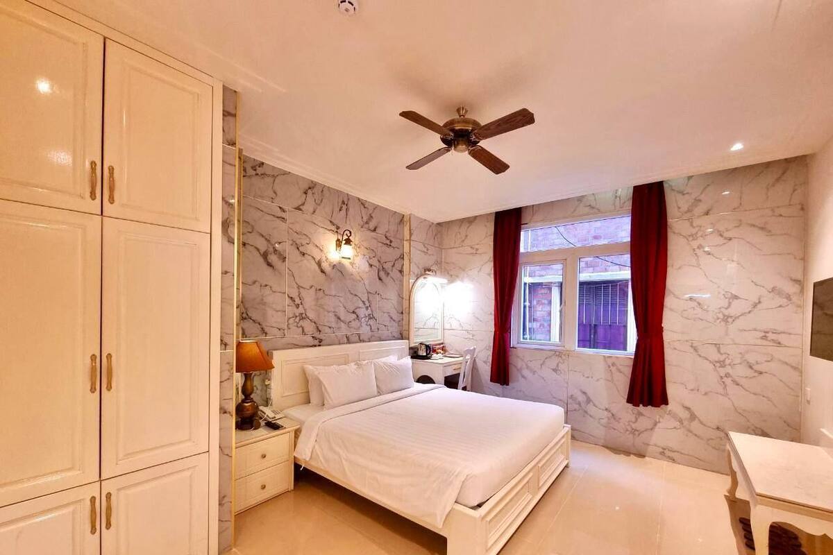 A bedroom with a bed, nightstands, a desk, a ceiling fan, and marble walls.
