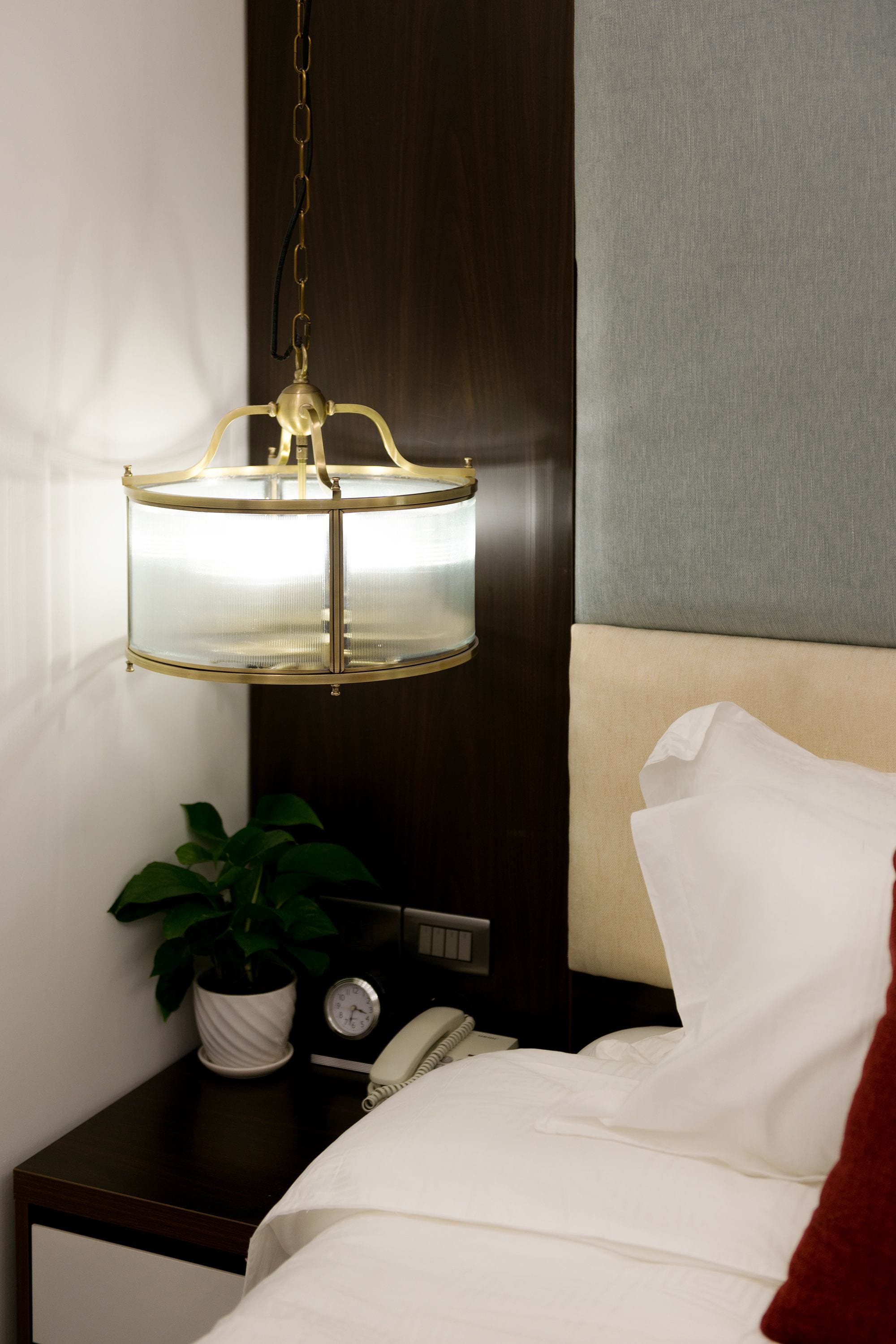 A hotel room with a bed, a bedside table with a plant, a clock, and a hanging lamp.