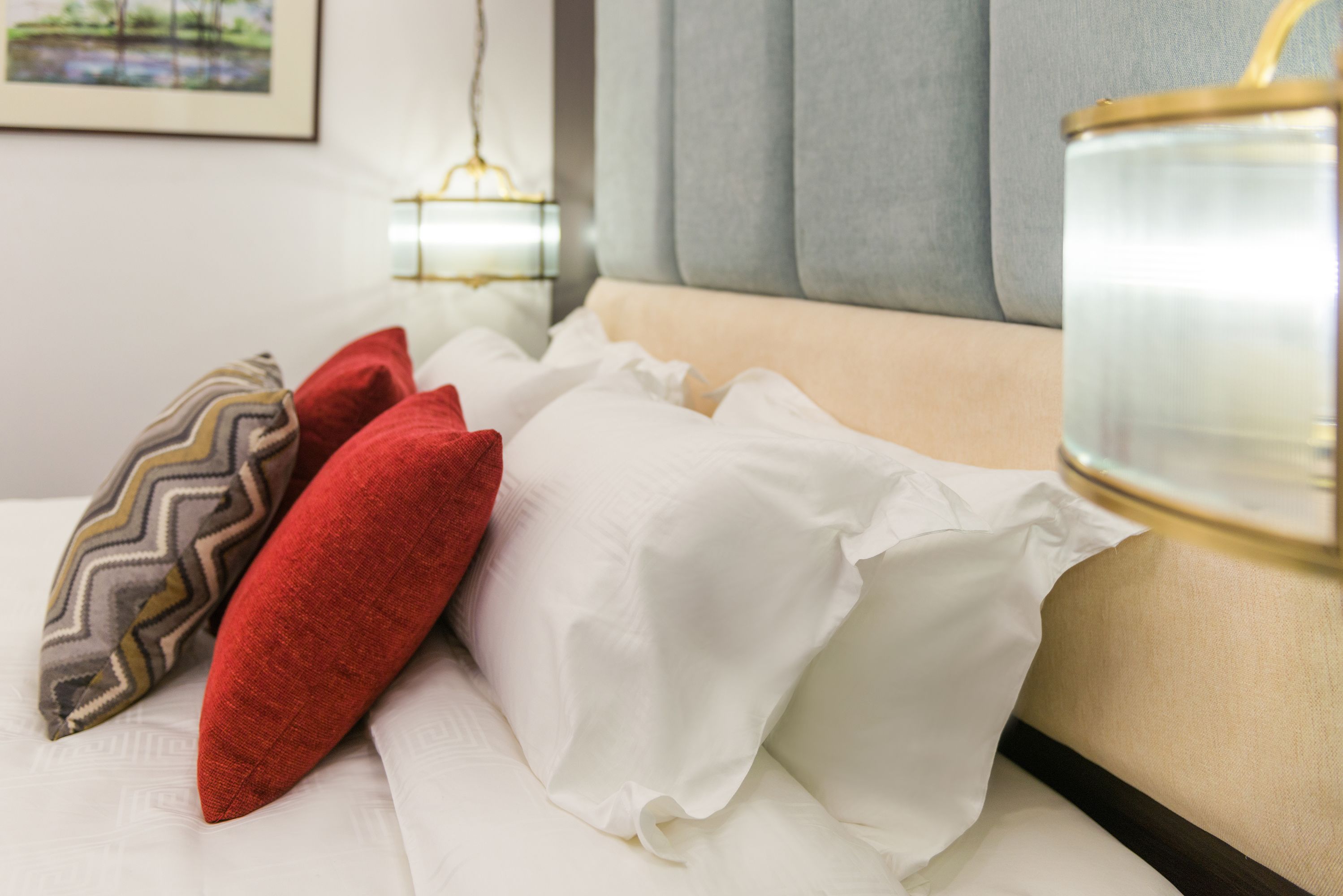 A neatly made bed with white and red pillows, a beige headboard, and a bedside table with a glass container and a lamp.