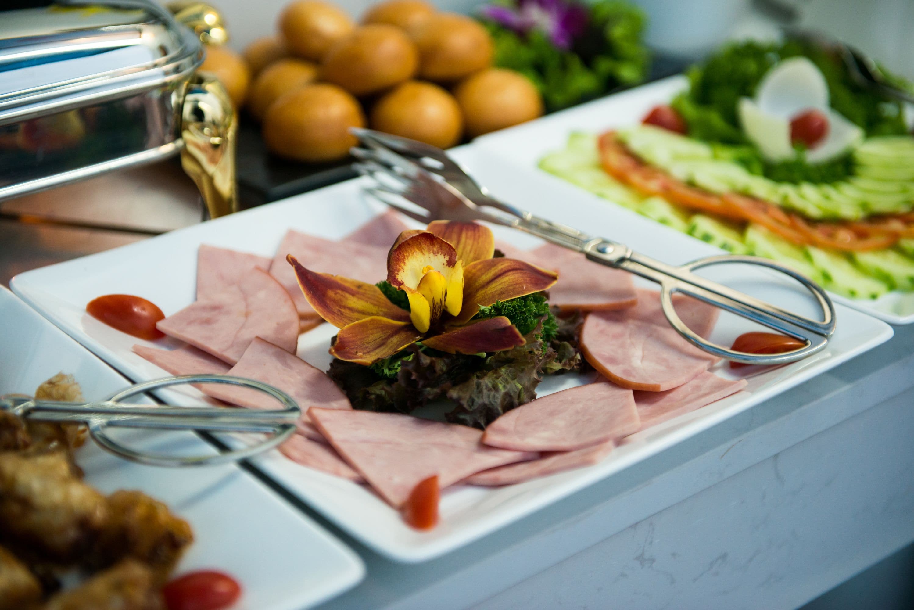 Un tavolo da buffet con diversi piatti, tra cui un elemento centrale con fiori e fette di carne, e un'insalata con verdure e un oggetto decorativo.
