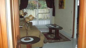 A living room with a sofa, a coffee table, and a small table with a decorative item. There is a window with curtains and a view of the outside.