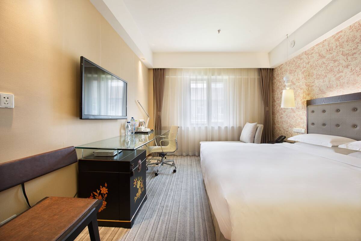 A hotel room with a bed, a desk with a computer, a chair, a TV, and a window with curtains.