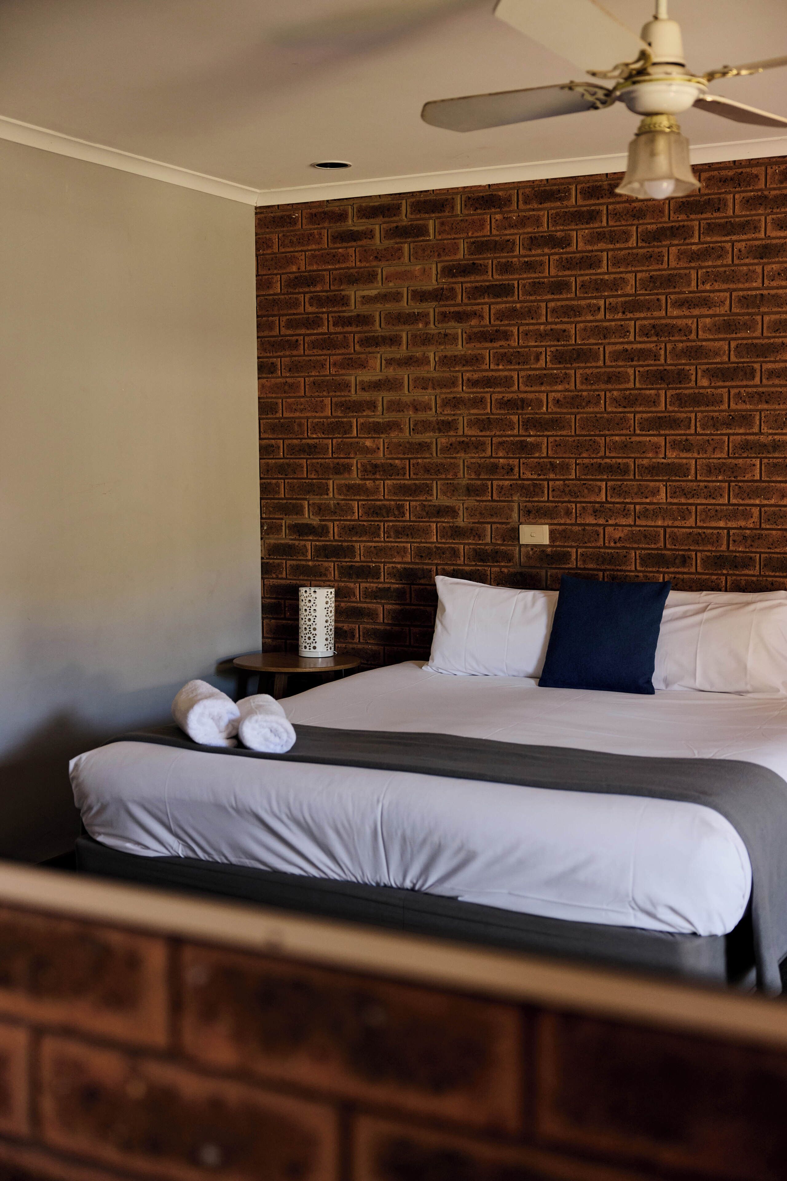 A hotel room with a brick-patterned wall, a ceiling fan, a bed with white bedding, and a small table with towels.