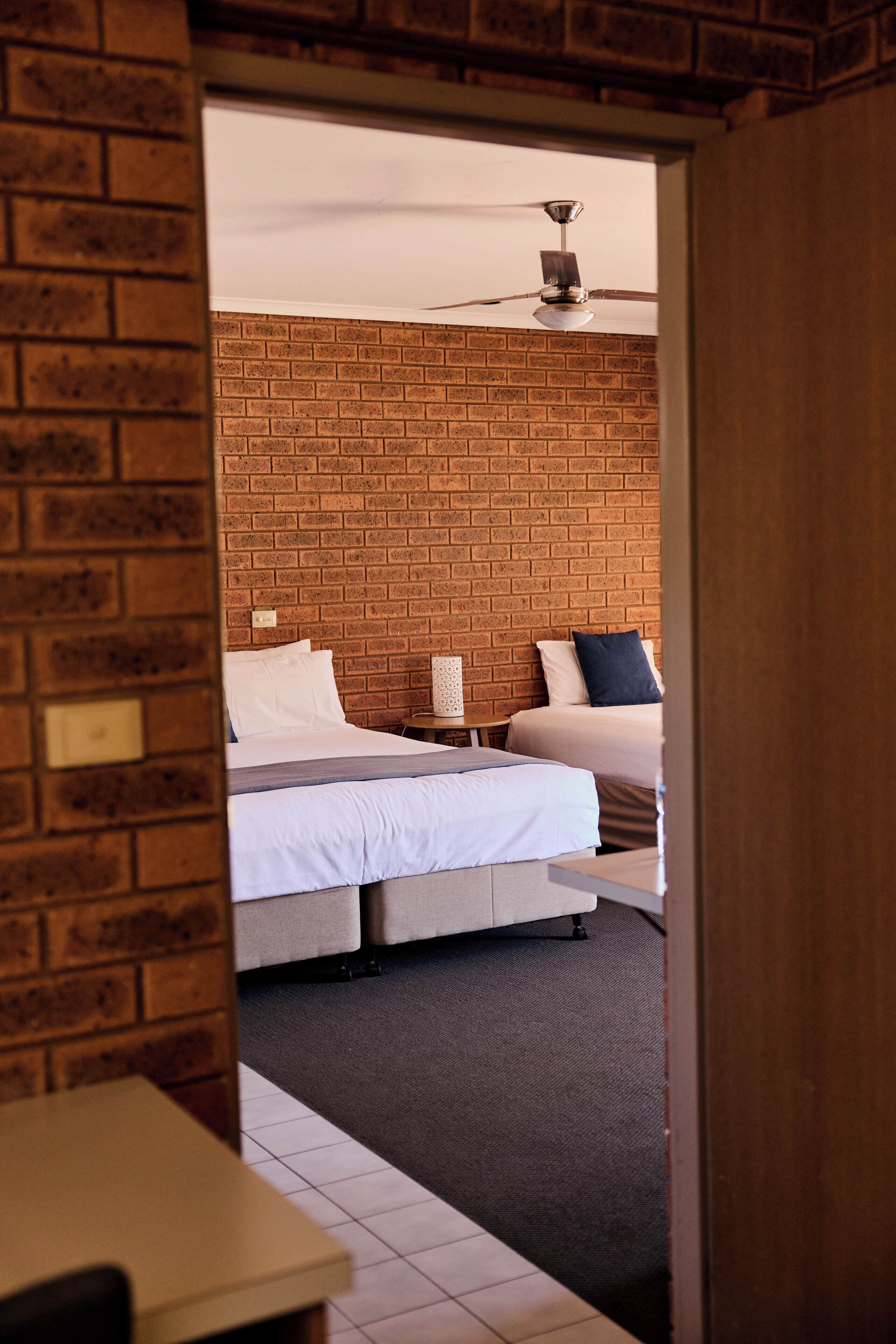 A hotel room with a brick wall, two beds, a ceiling fan, and a small table.