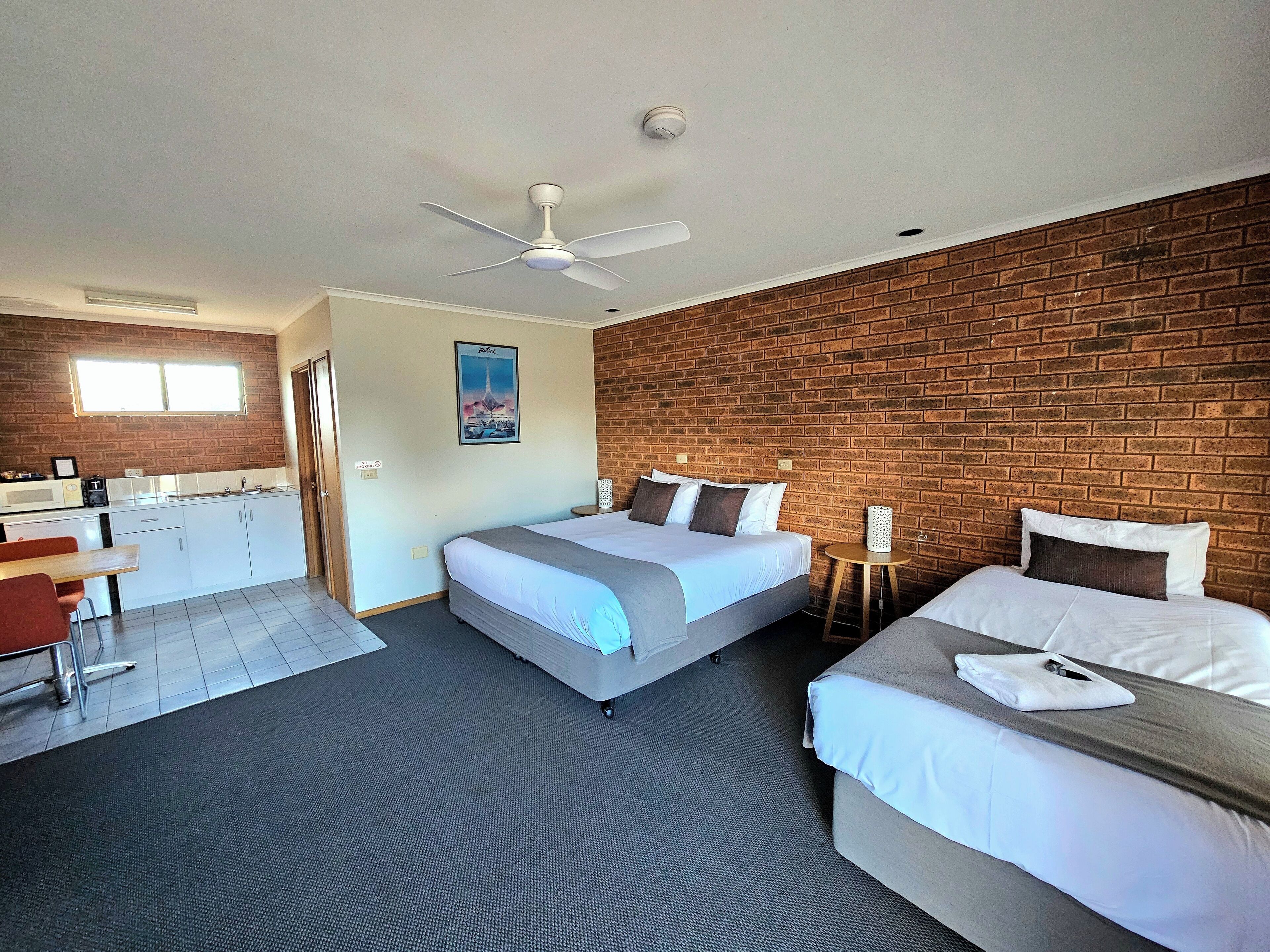 A hotel room with a bed, a desk, and a chair. There is a brick wall and a ceiling fan.