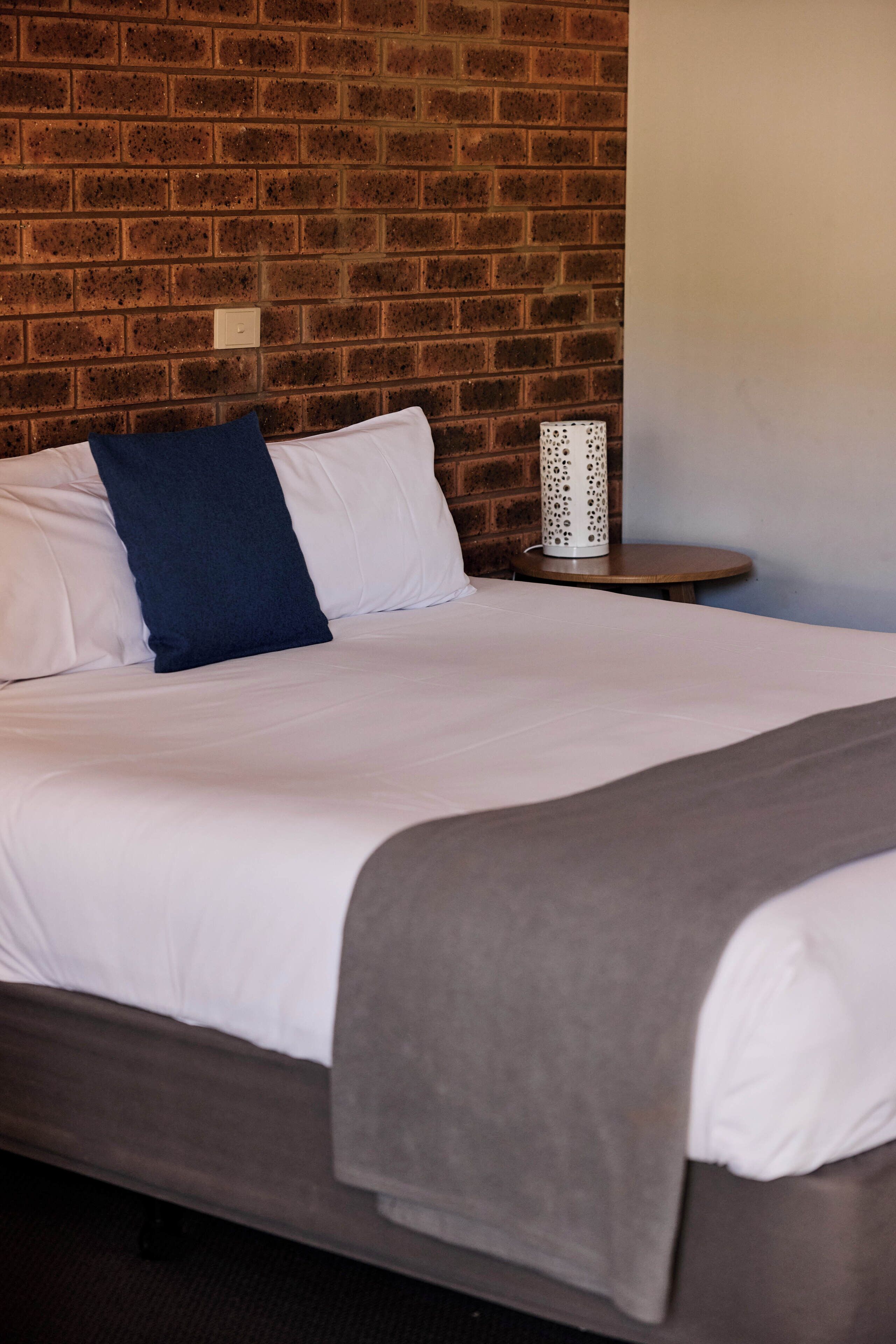 Uma cama com lençóis brancos, um travesseiro azul, um cobertor cinza e um abajur branco sobre uma mesa redonda ao lado da cama.