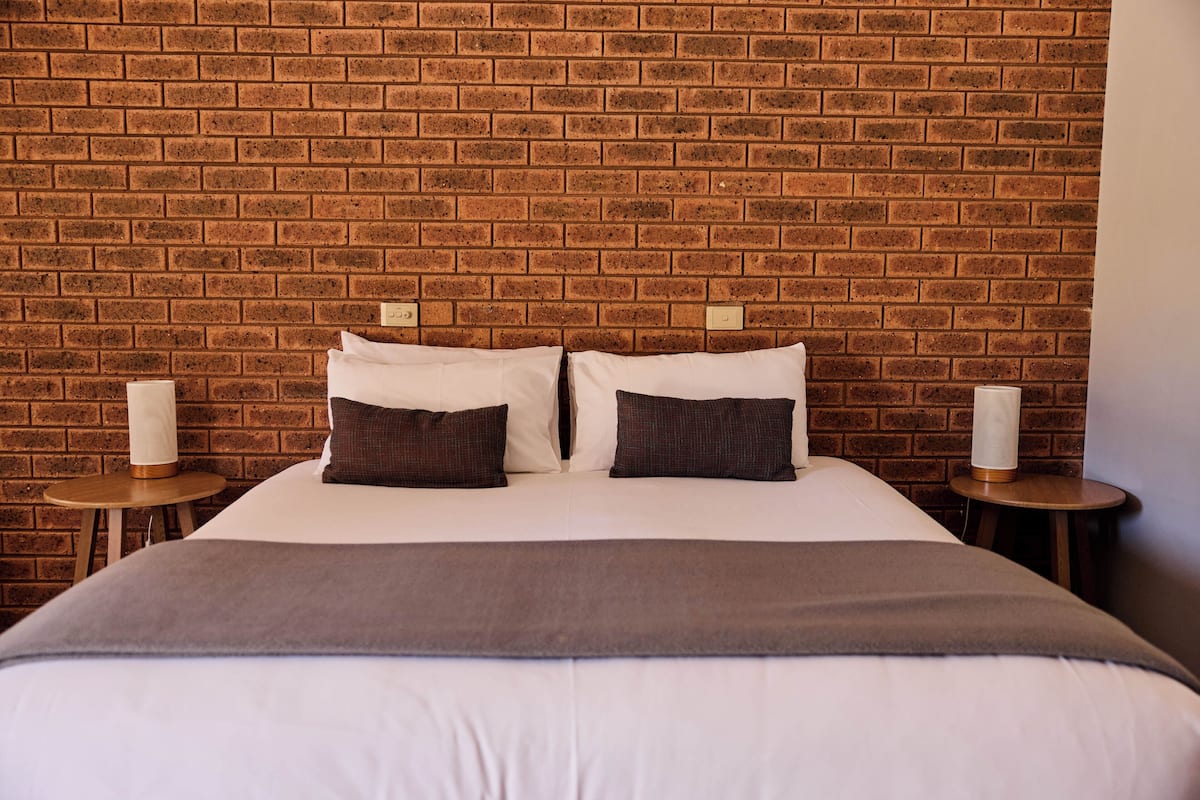 A hotel room with a brick wall, a bed with white bedding and two pillows, and two bedside tables with lamps.