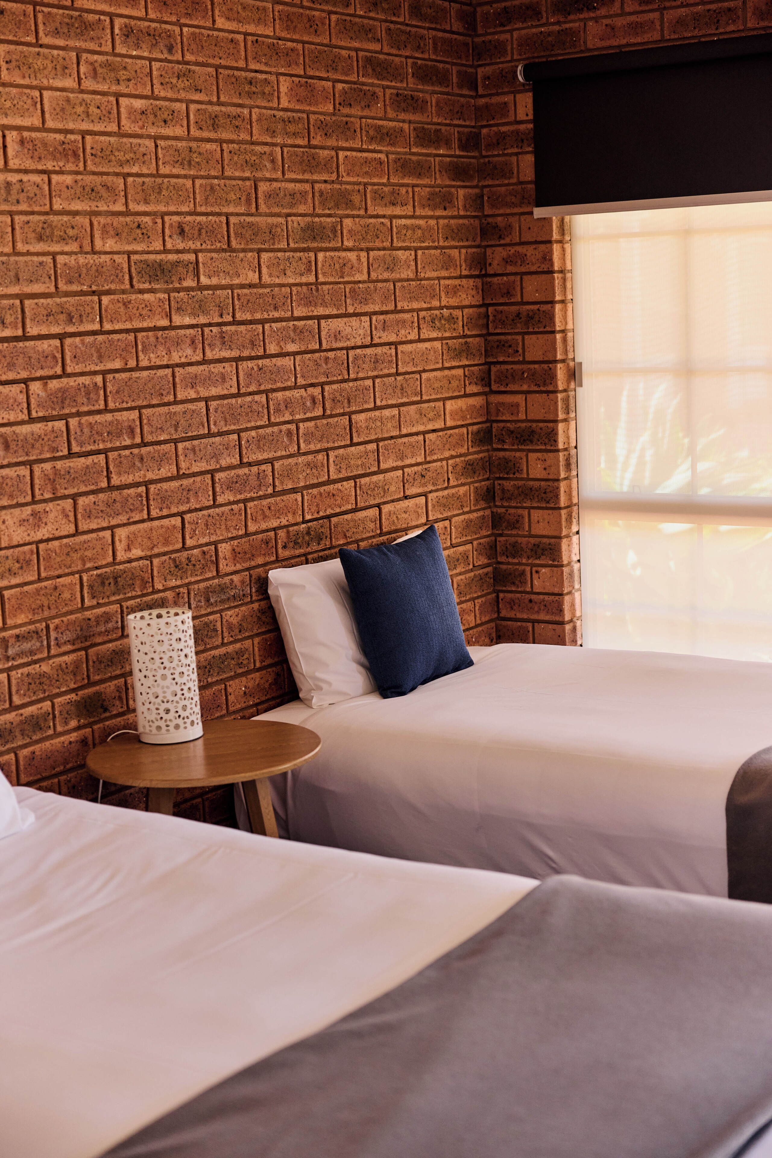A hotel room with a brick wall, two beds, a small round table, and a window with blinds.
