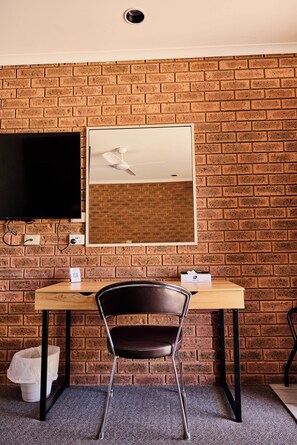 Une pièce avec un mur en briques, un téléviseur à écran plat fixé au mur, un bureau en bois avec une chaise et une poubelle blanche.