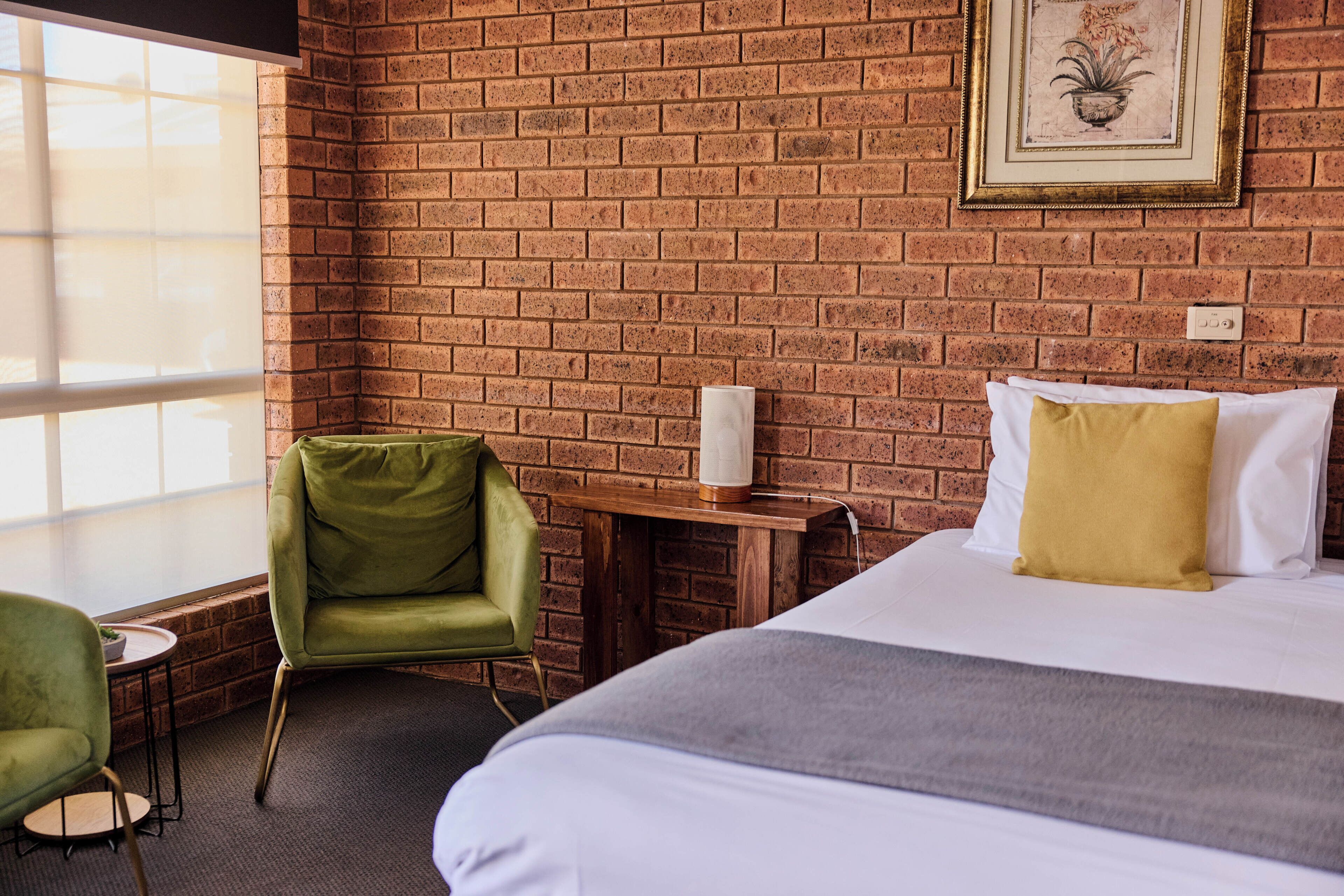 A hotel room with a brick wall, a bed with white and yellow pillows, a green armchair, and a small table with a lamp.