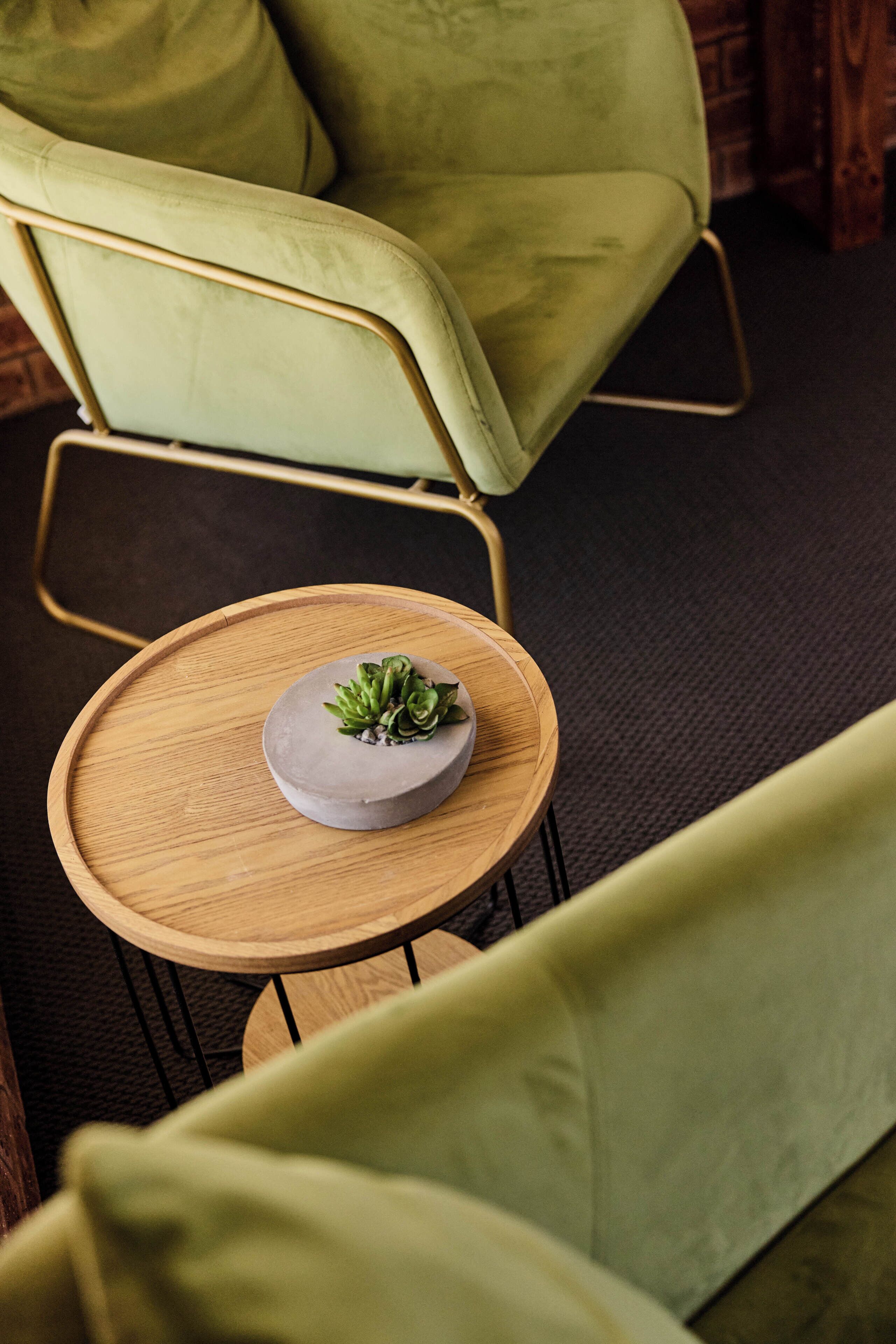 A green upholstered chair, a small round wooden table with a gray ceramic bowl and green plants, and a dark carpeted floor.