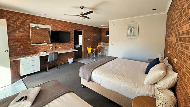 A hotel room with a brick wall, a bed, a desk with a chair, a TV, and a ceiling fan. - Bridge Motor Inn (Tocumwal)