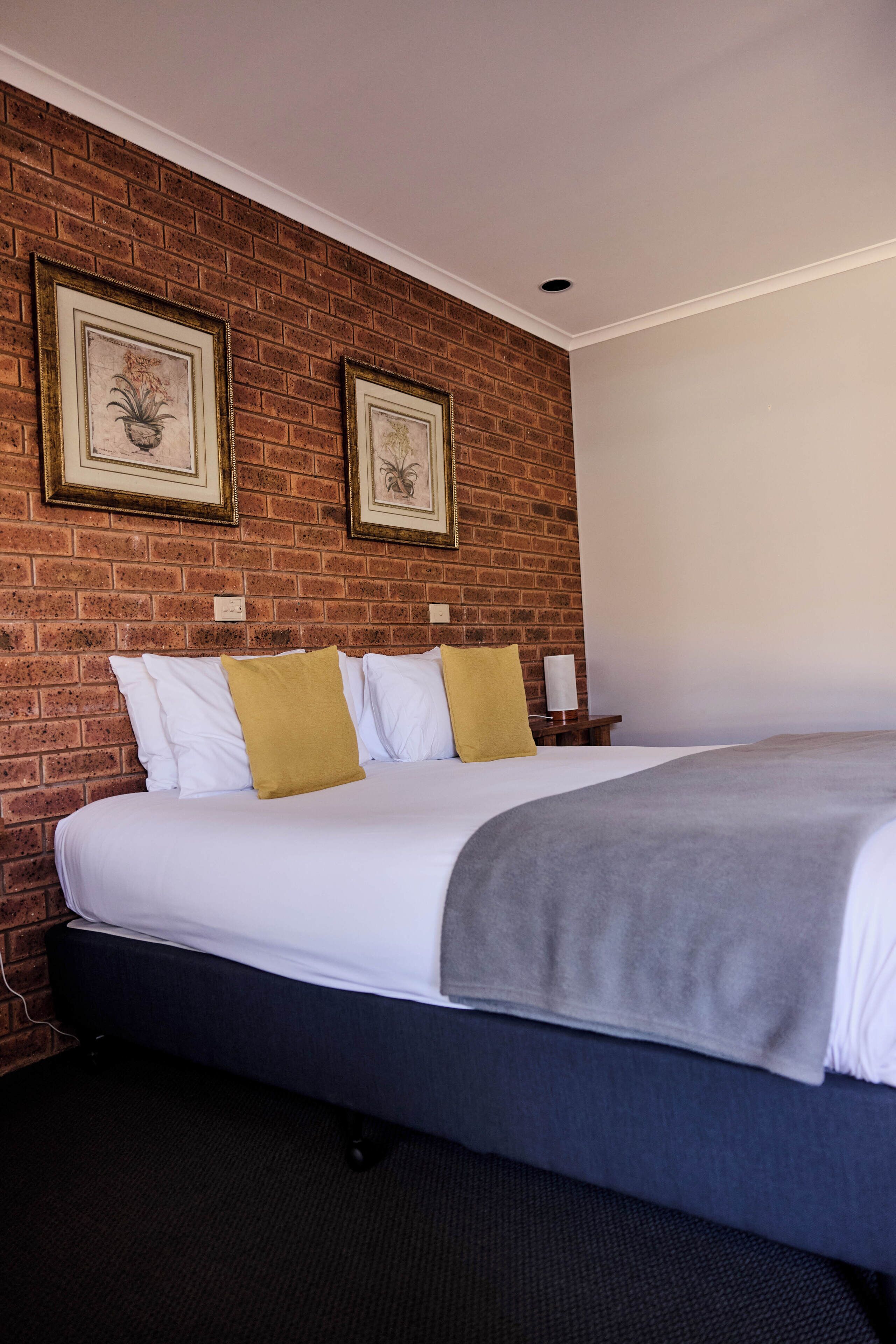 Una habitación de hotel con una pared de ladrillo, una cama con ropa blanca y una manta gris, dos cuadros en la pared y una mesita de noche con una lámpara.