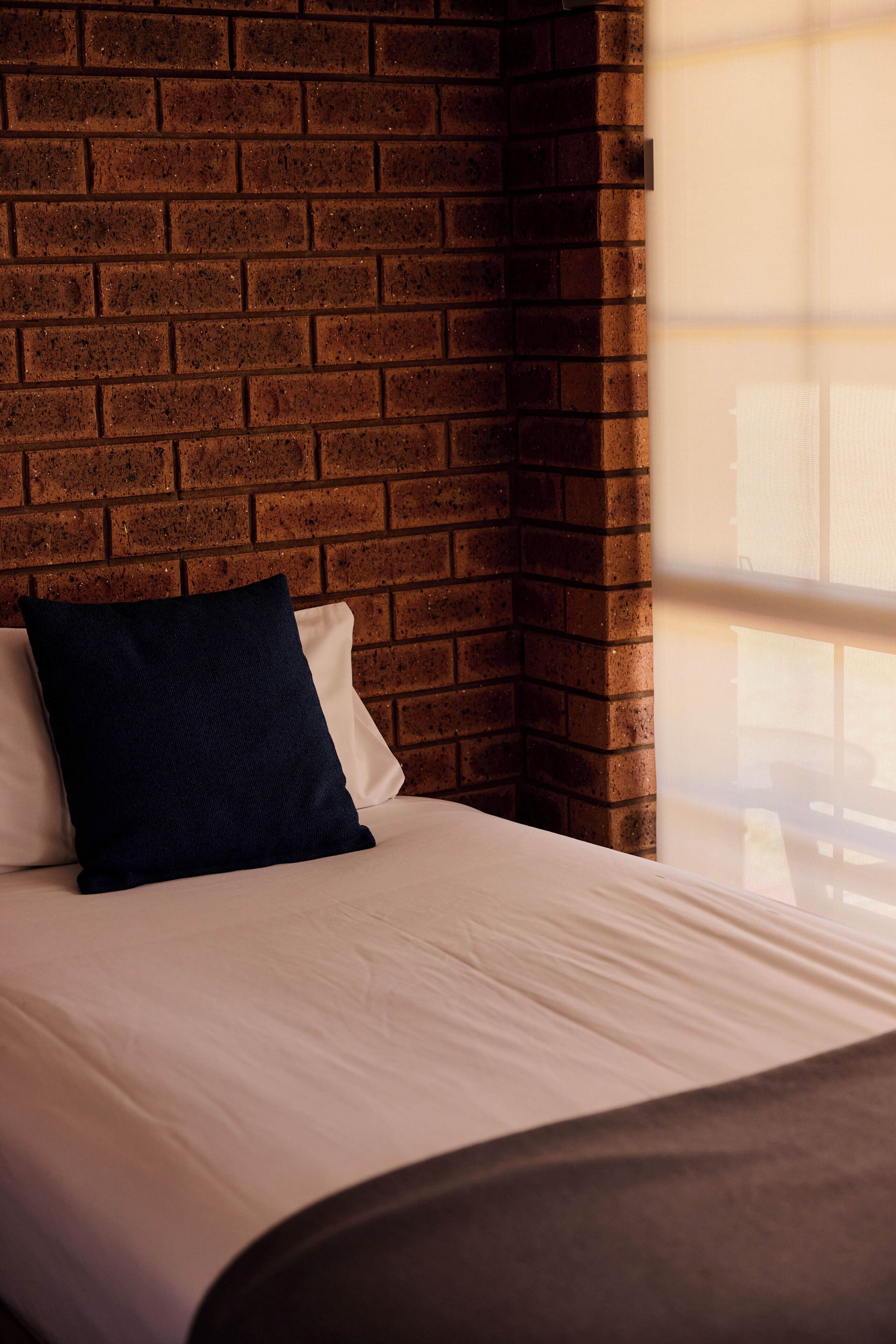 Cama com roupa de cama branca e travesseiro escuro, com parede de tijolos ao fundo.