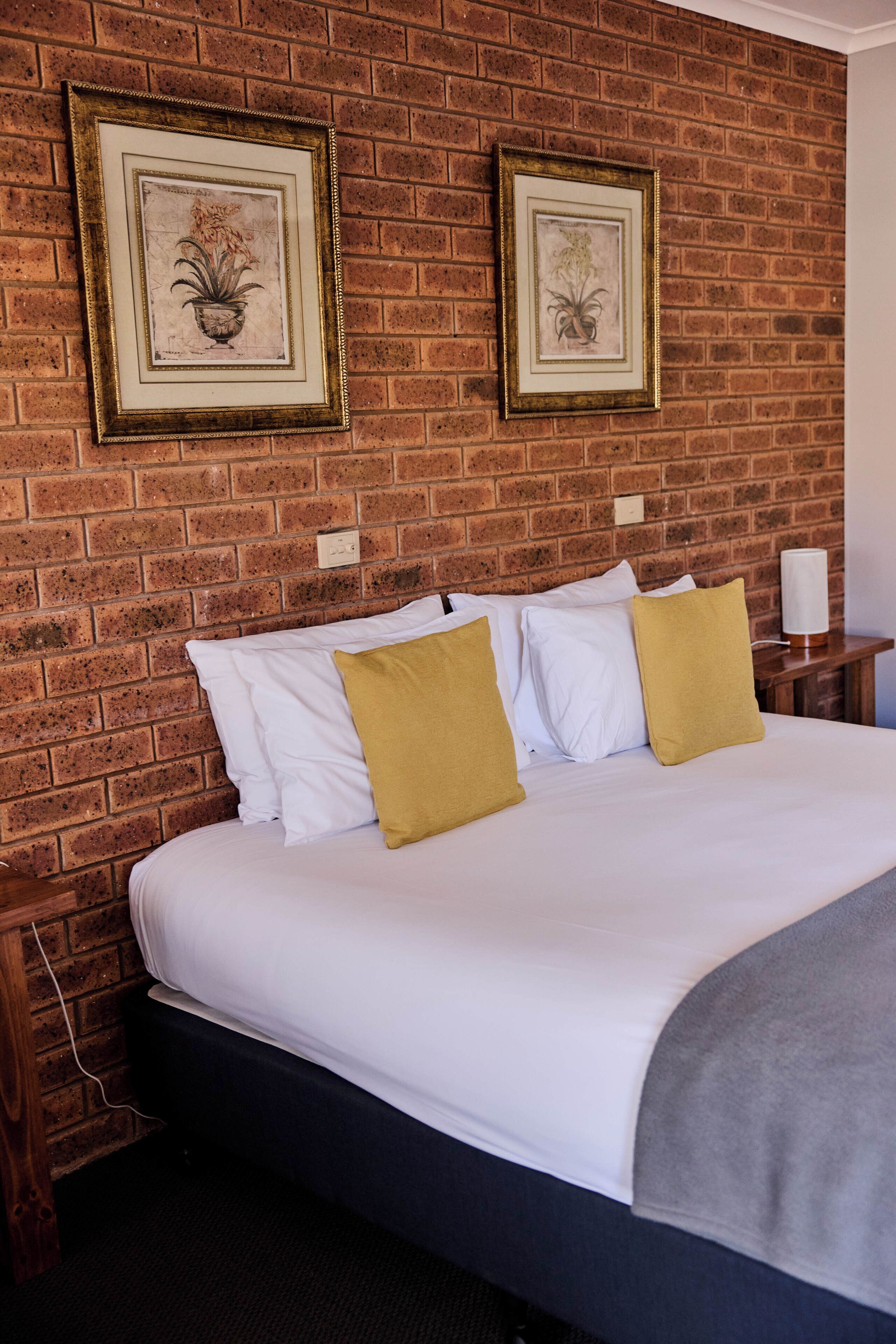 Une chambre d’hôtel avec un mur en briques, un lit avec des oreillers blancs et moutarde, et deux tableaux encadrés accrochés au mur.