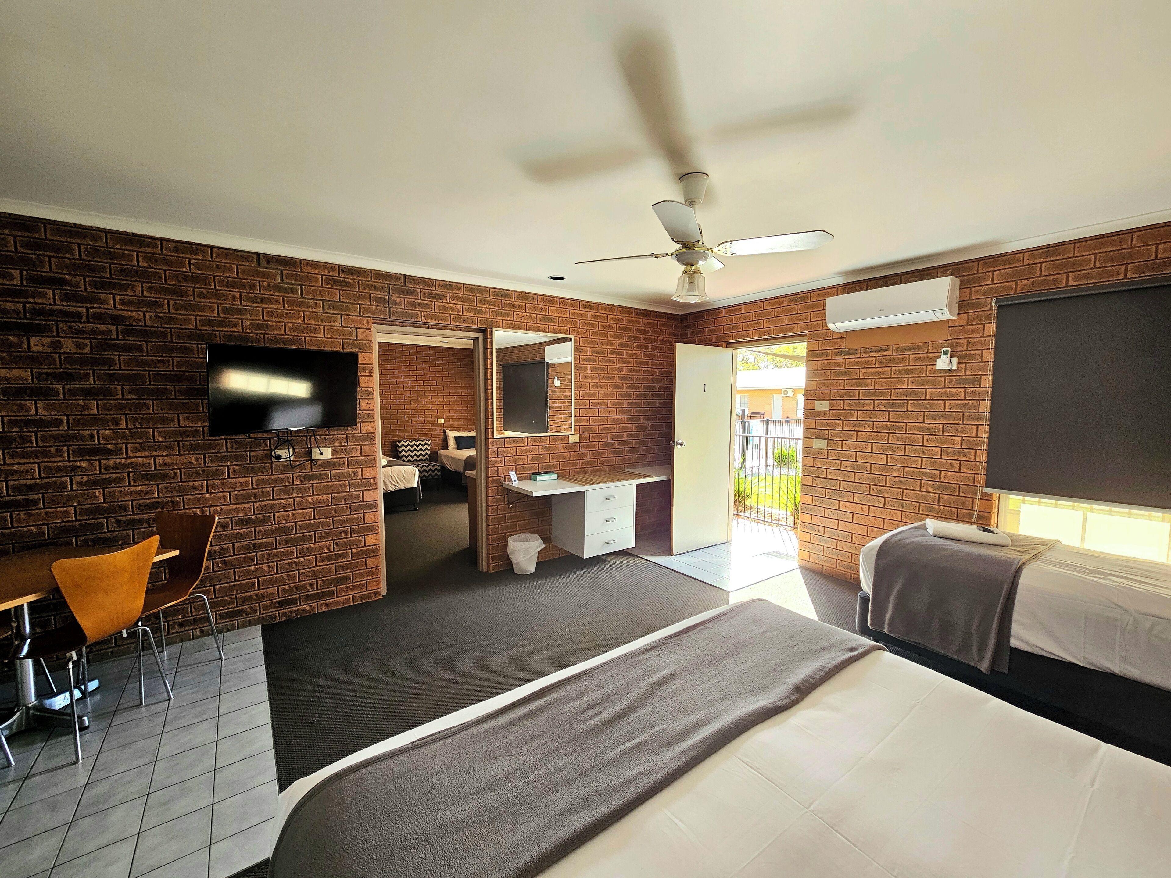 A hotel room with brick walls, a ceiling fan, a TV, a desk with a chair, a bed, and a window.