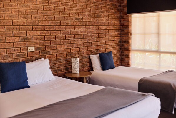Une chambre d’hôtel avec deux lits, un mur en briques, une petite table avec une lampe et une fenêtre avec des stores.
