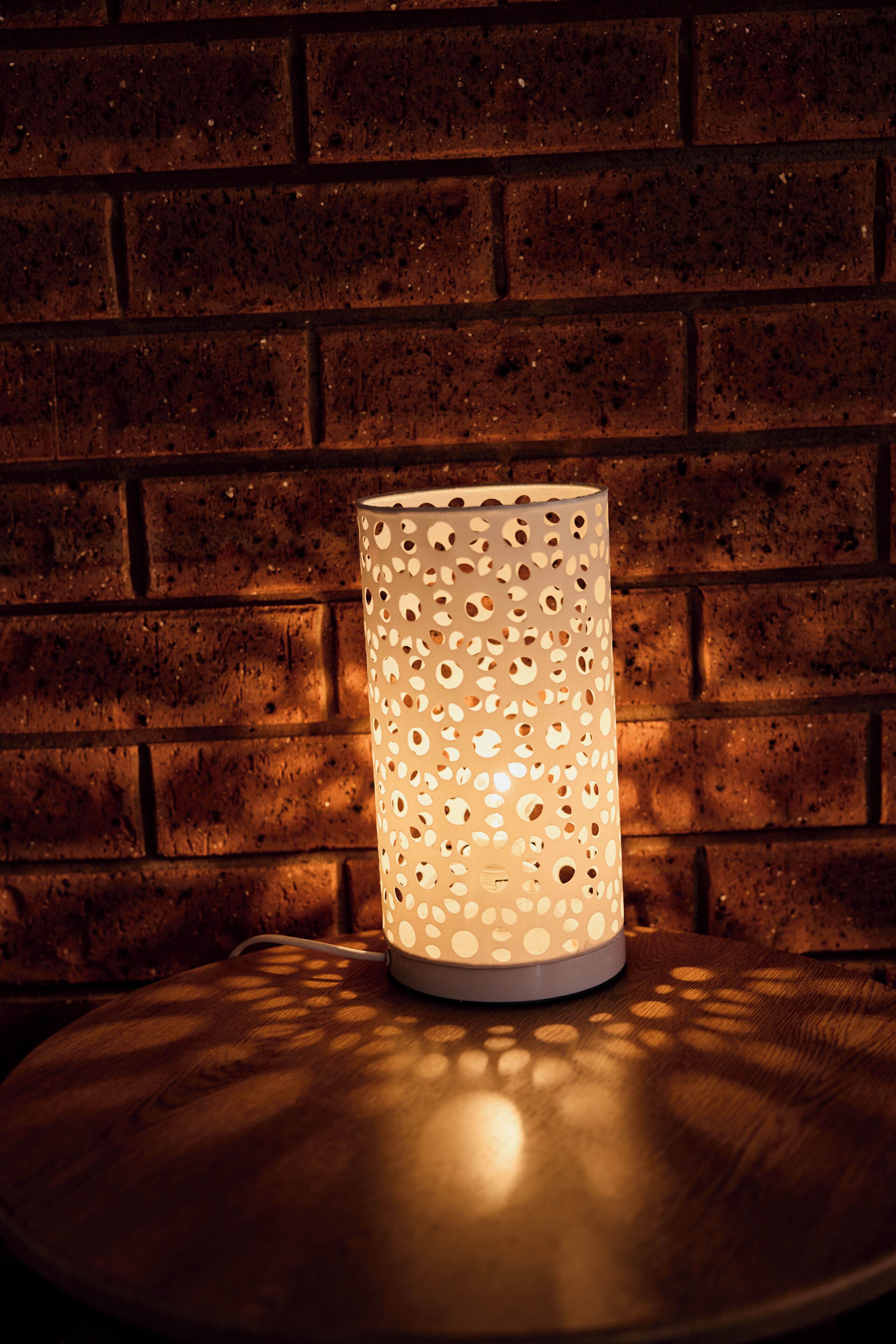 A cylindrical lamp with a perforated design on a round table against a brick wall.