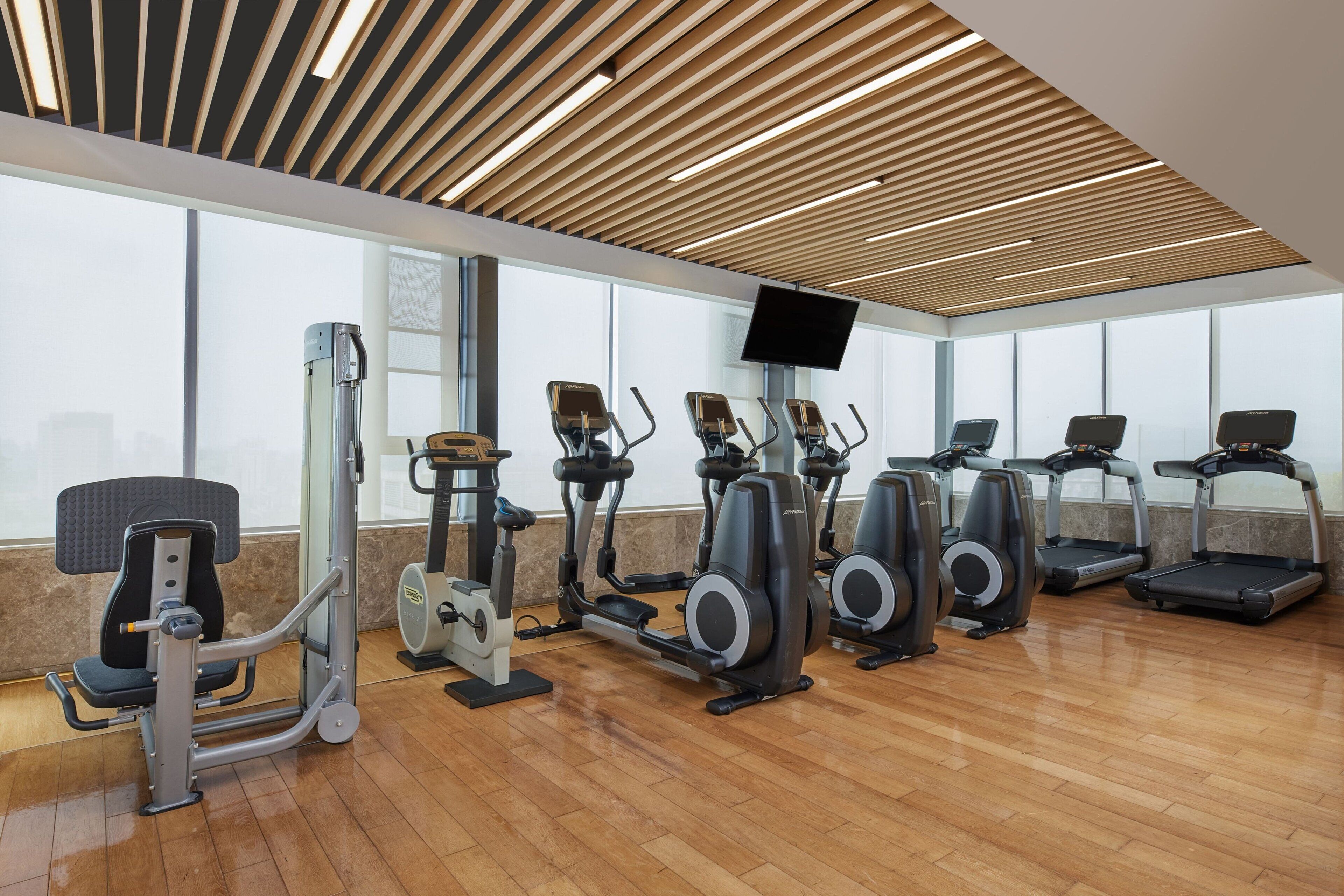 A gym with various exercise machines, a television mounted on the wall, and large windows providing natural light.