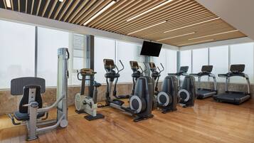 A gym with various exercise machines, a television mounted on the wall, and large windows providing natural light.