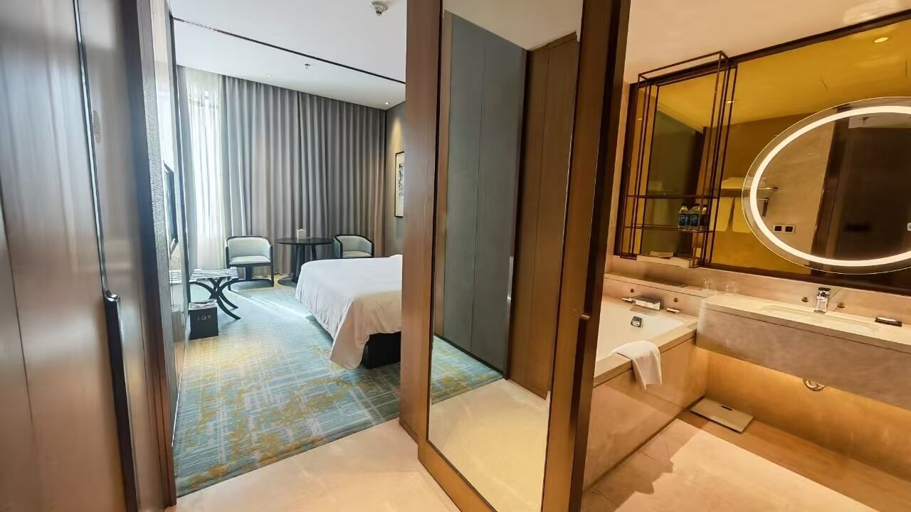 A modern hotel room with a large bed, a separate sitting area with a table and chairs, and a bathroom with a large mirror and sink.